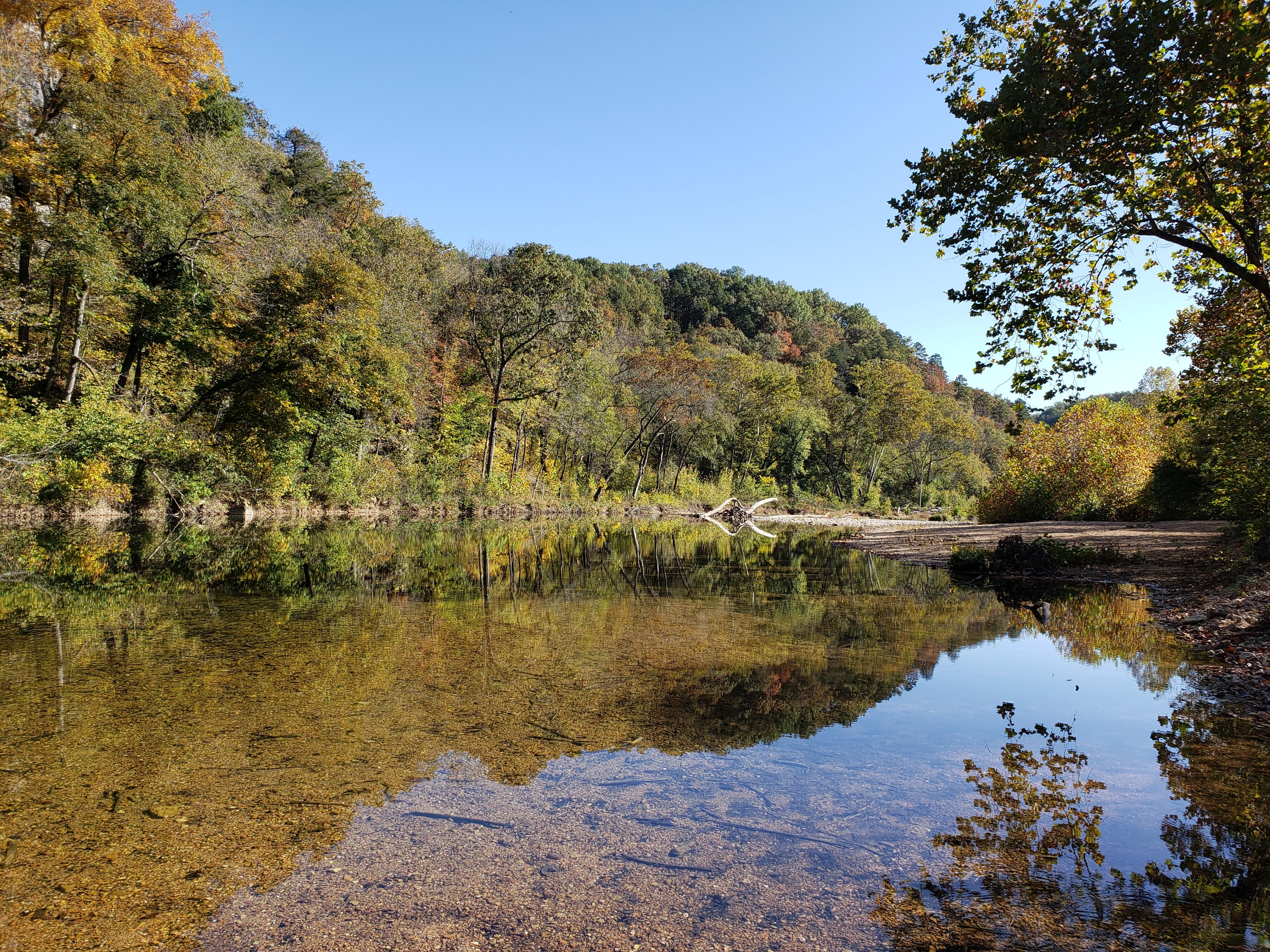 A shallow river reflects trees and mountains like a mirror.