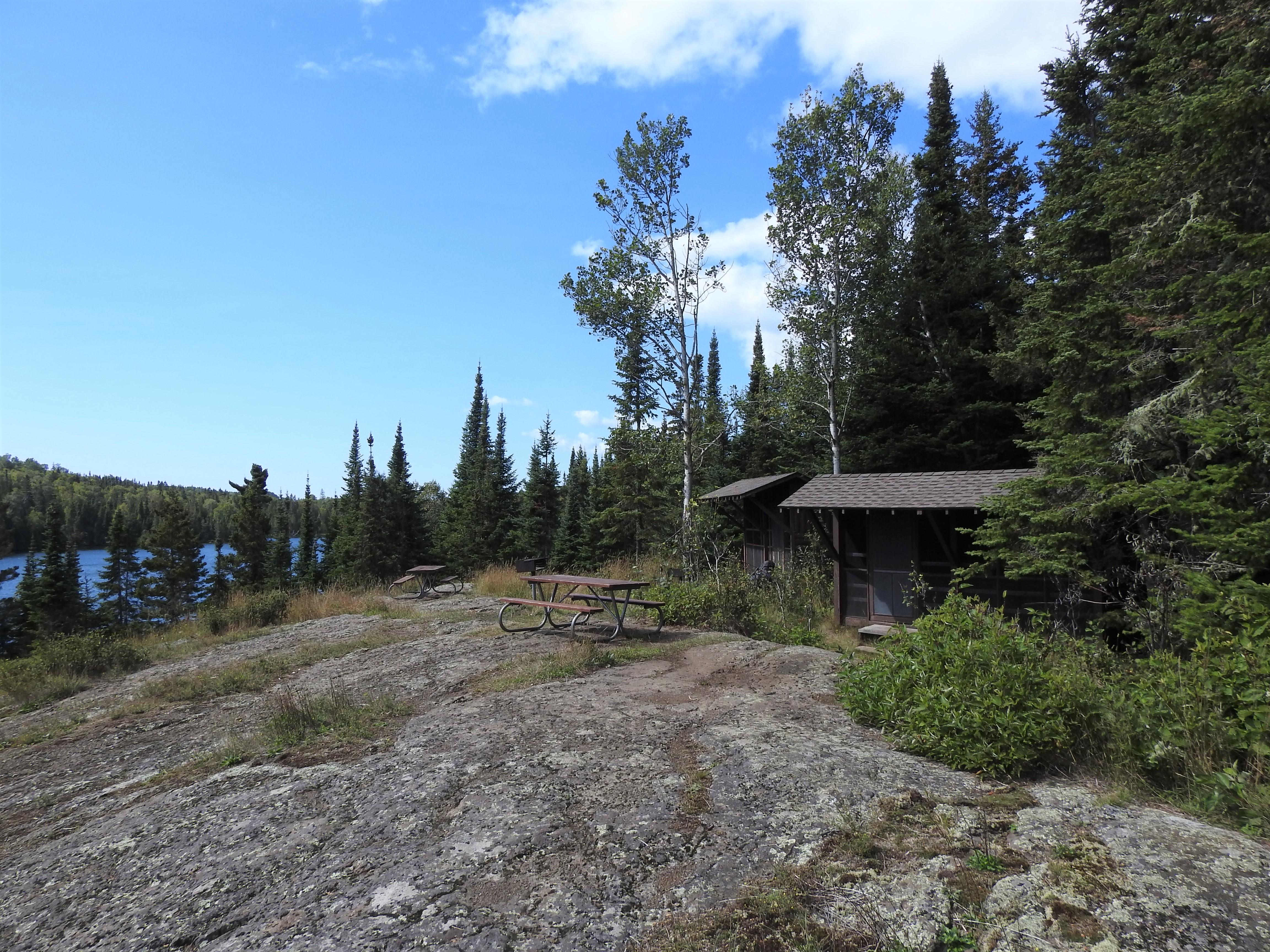 Rocky shoreline with two shelters along the treeline with blue sky overhead.