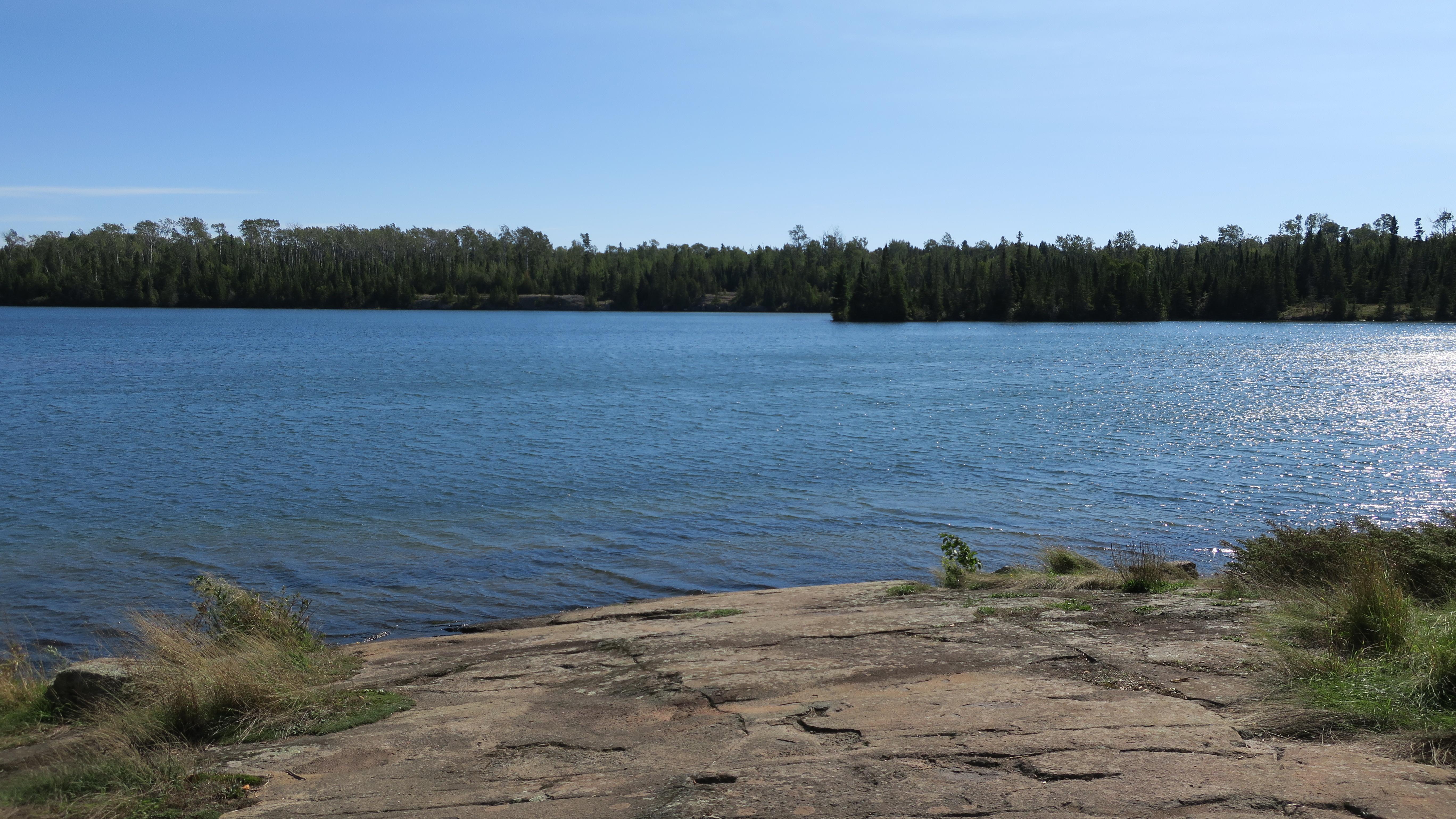 Blue sky, calm water, rock shoreline in foreground.