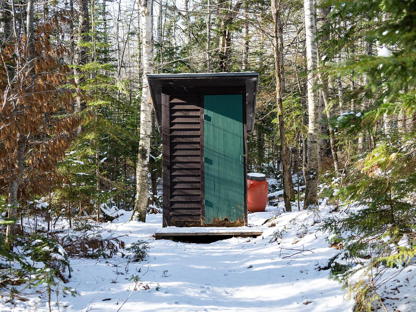 A snowy trail leads to a wooden pit toilet tucked in the woods.