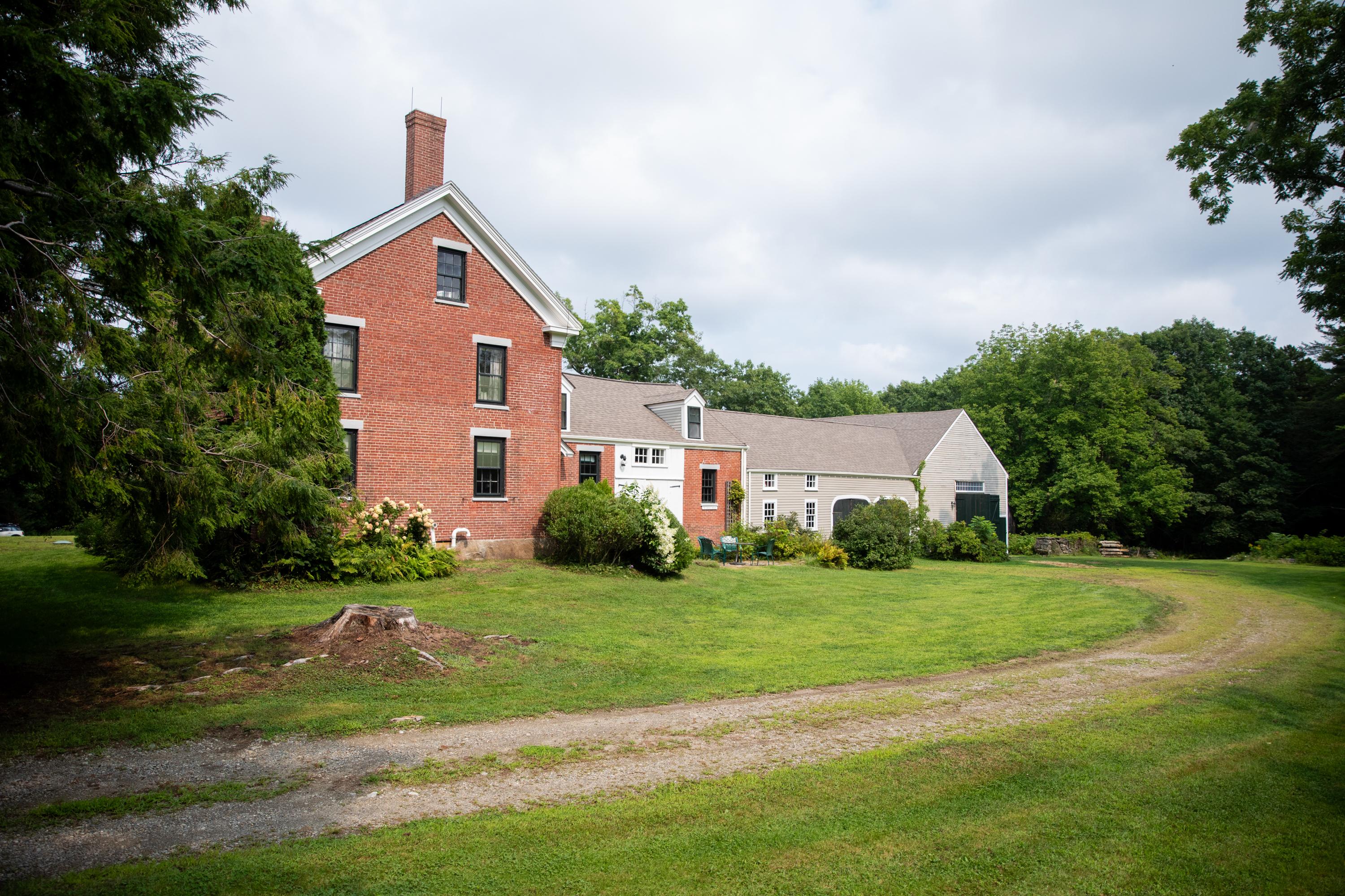 A sandy brown driveway curves through green grass towards the two story-red brick homestead and grey