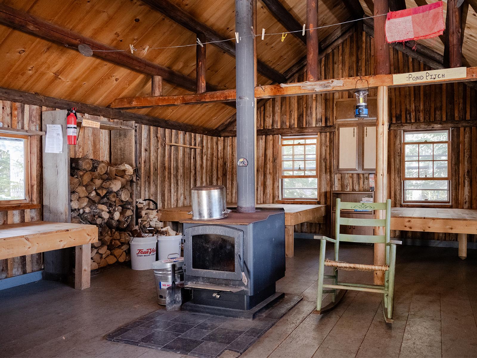 Woodstove in the middle of a log cabin. Wood stacked for use against the wall and wood bunks behind.