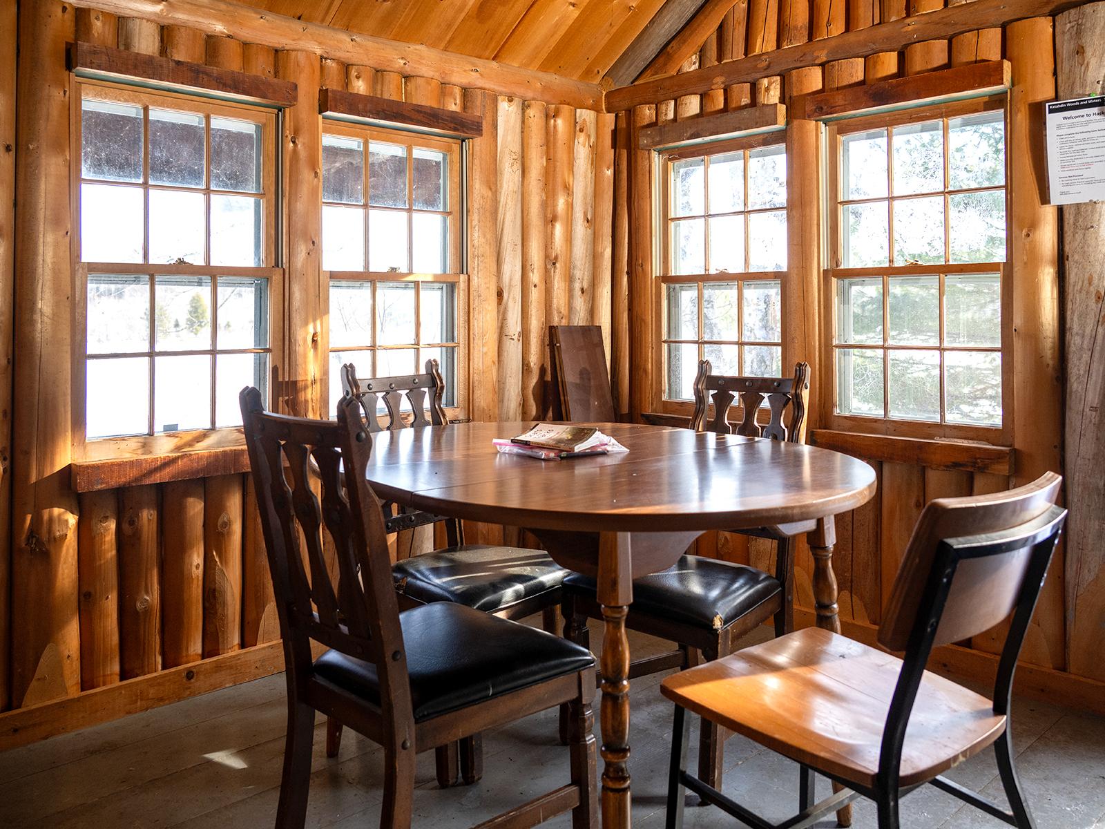 A wooden dining table with 4 chair around it inside a wood cabin. 4 slim windows let light inside.