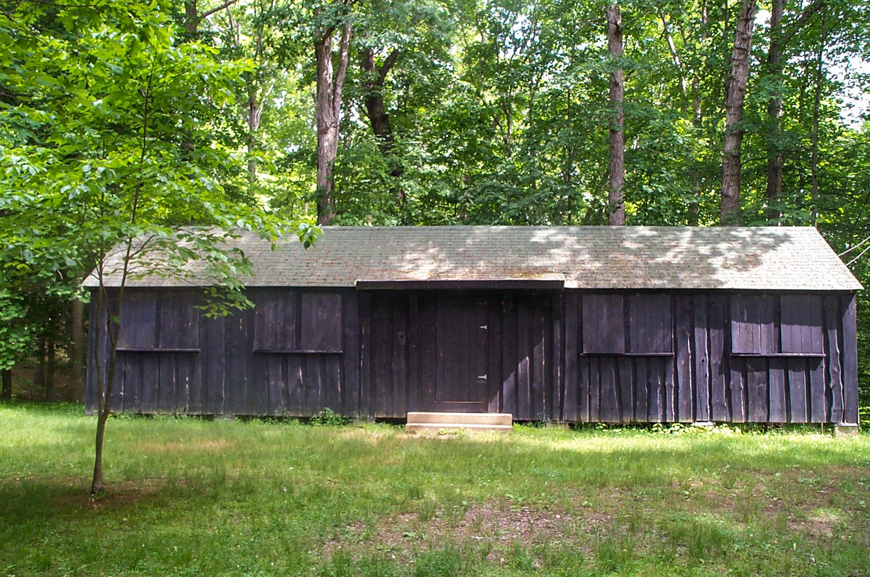 A dark brown, rectangular wooden cabin sits among green trees with a grassy front lawn