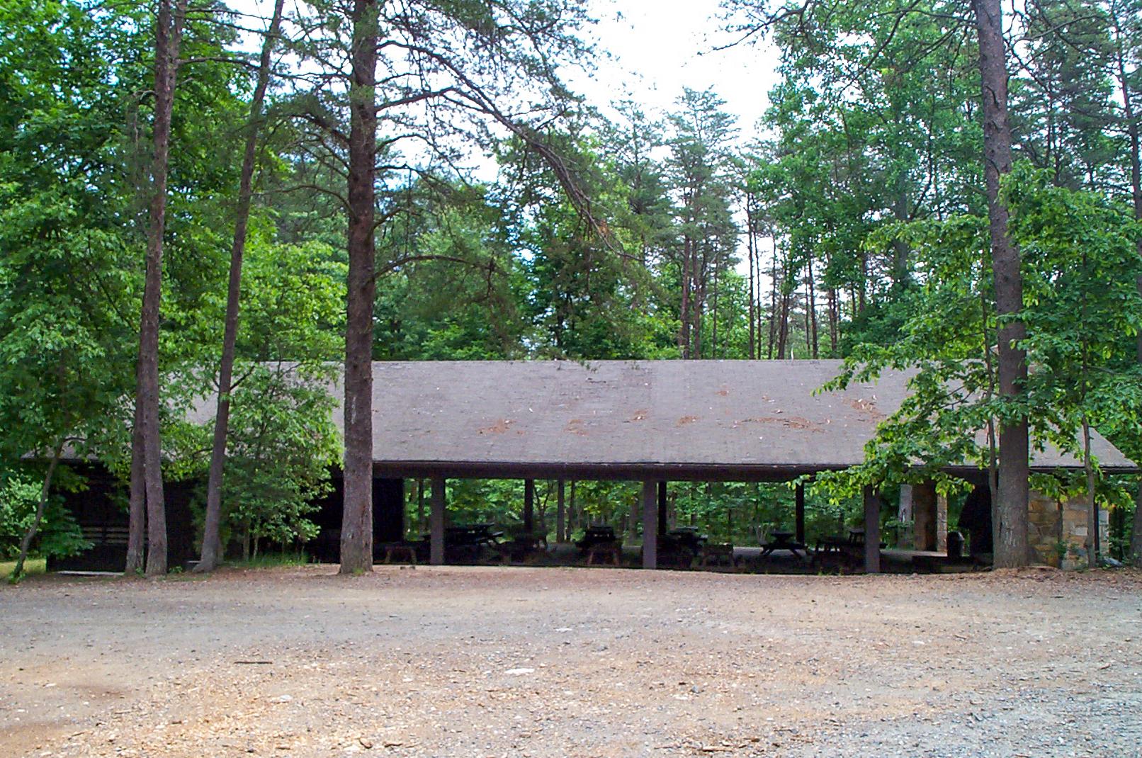 A wooden pavilion shelters several wooden picnic tables. Green trees surround the pavilion
