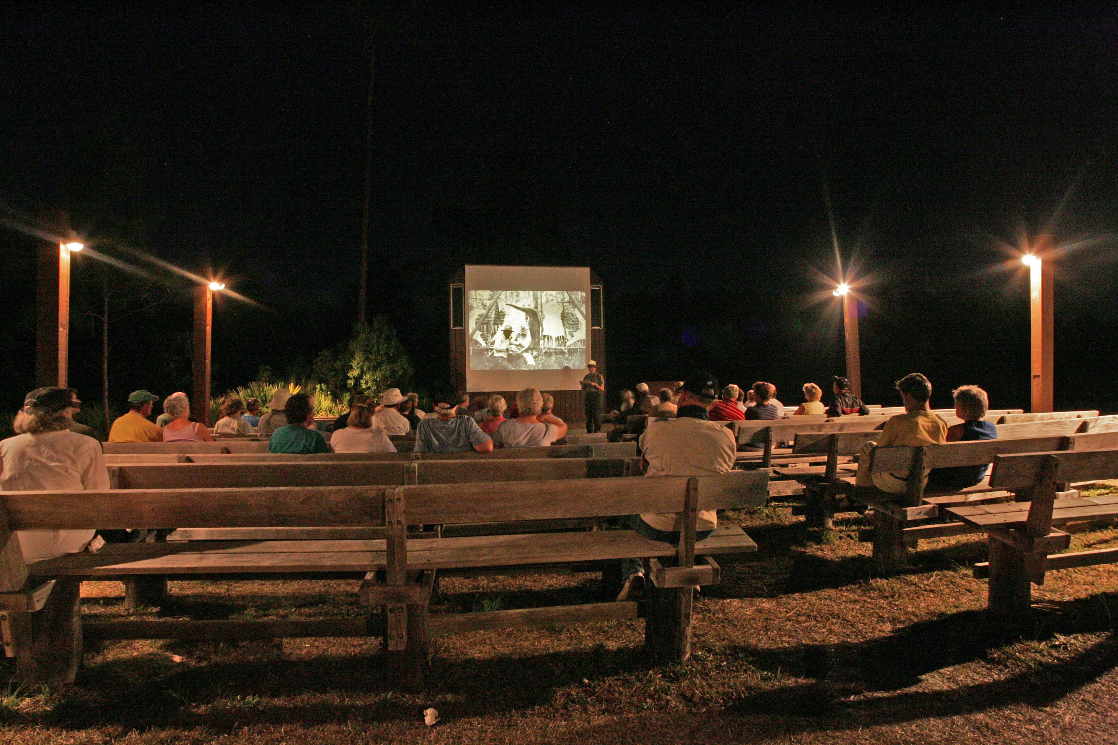 People sit on wooden benches listening to a Park Ranger deliver a program. It is night time.