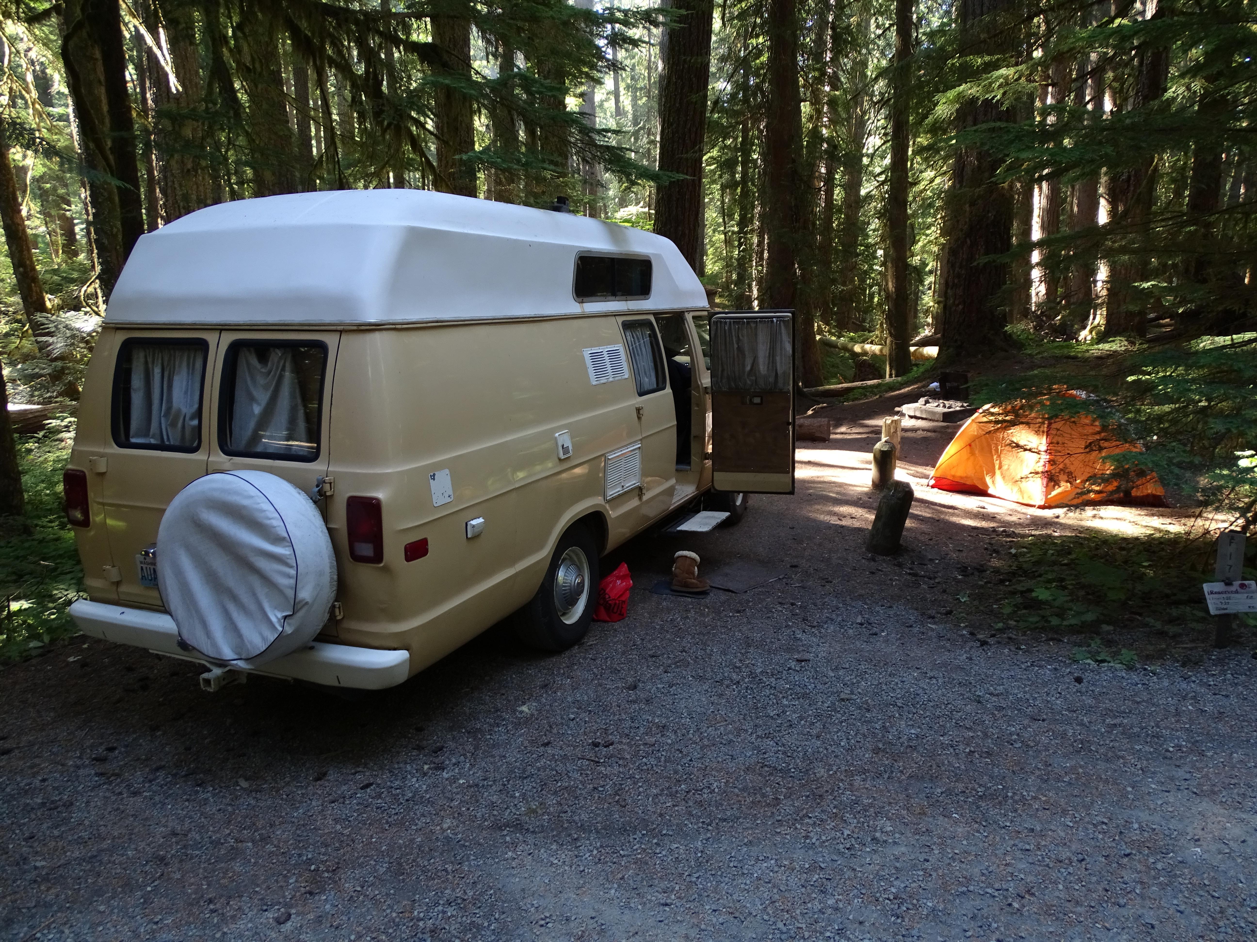 A tan and white van next to an orange tent in a forested campsite.