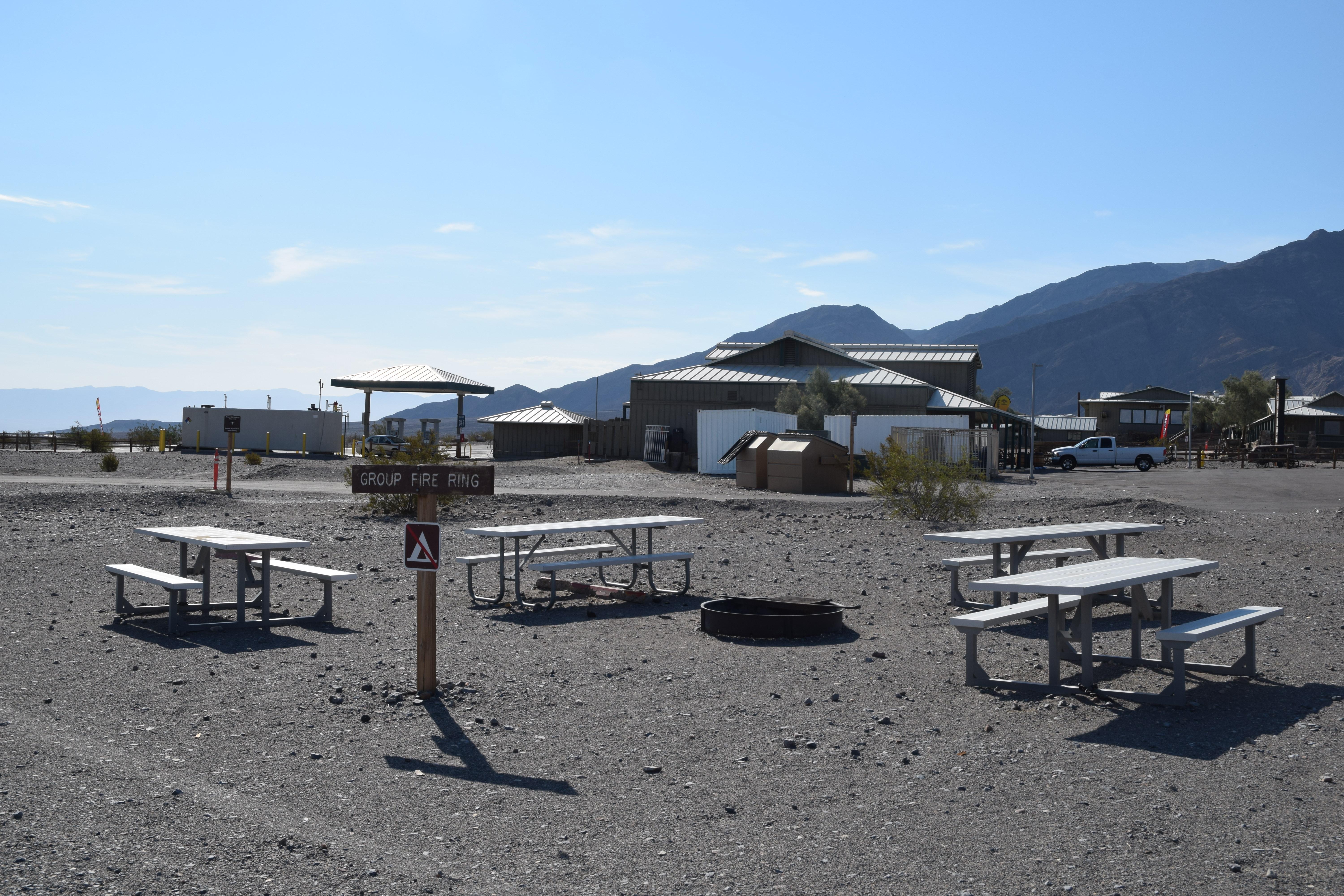 4 metal picnic tables surround a metal fire ring on an open, gravel area.