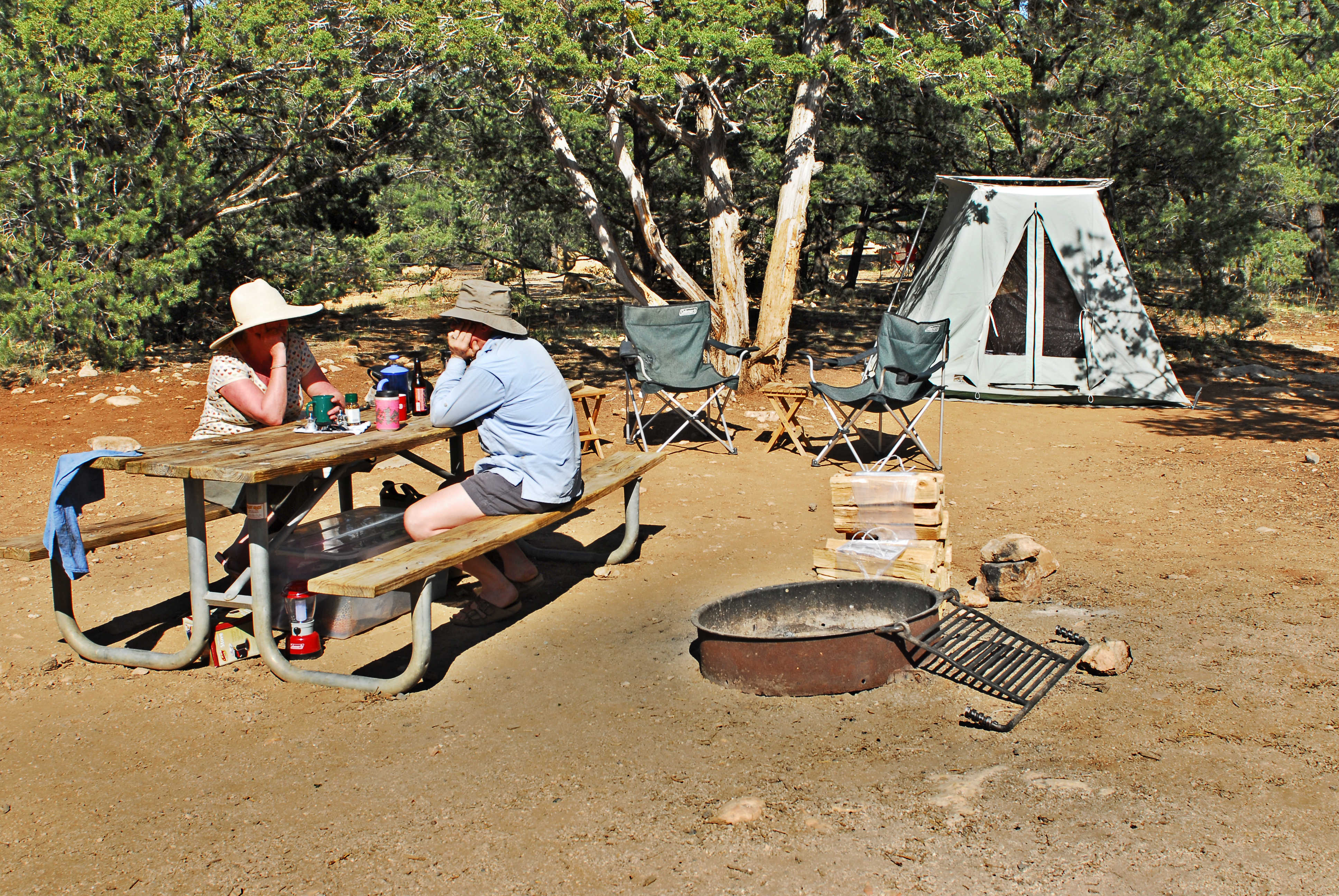 A man and a woman sit at a picnic table in the sunlight a tent and chairs surround a firepit