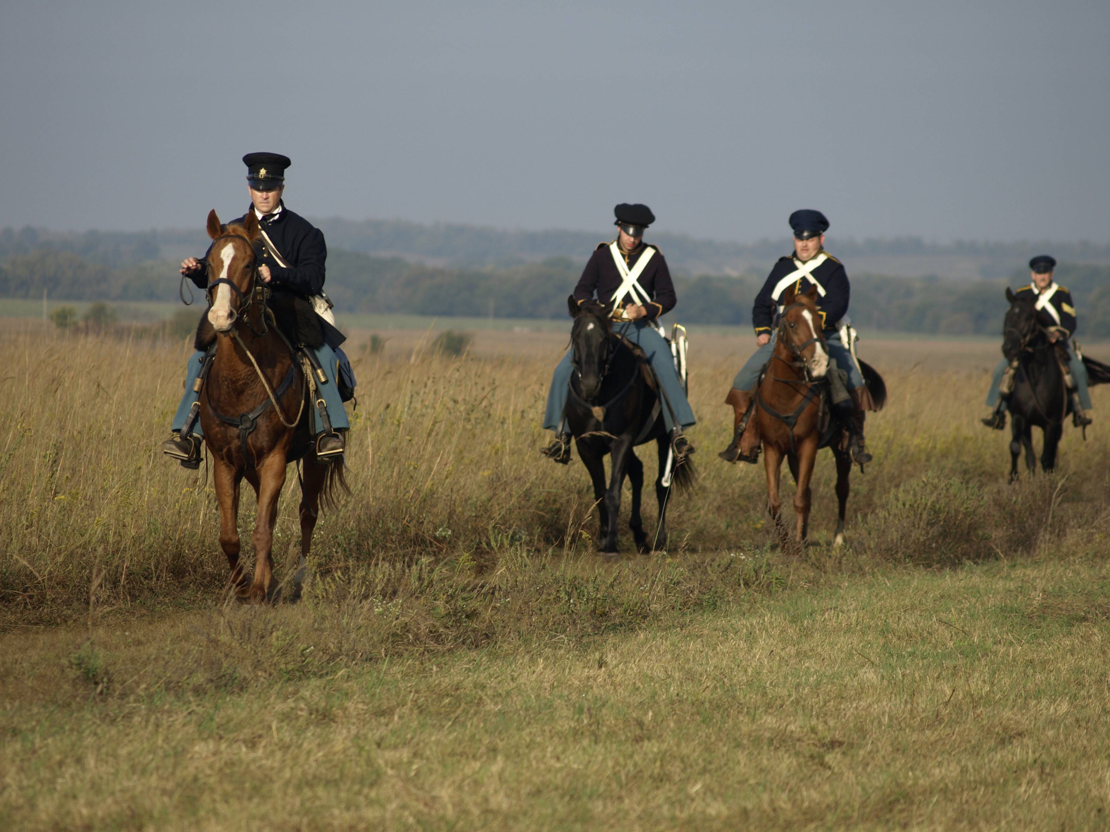 Four soldiers on horseback in a line riding through prairie grass.