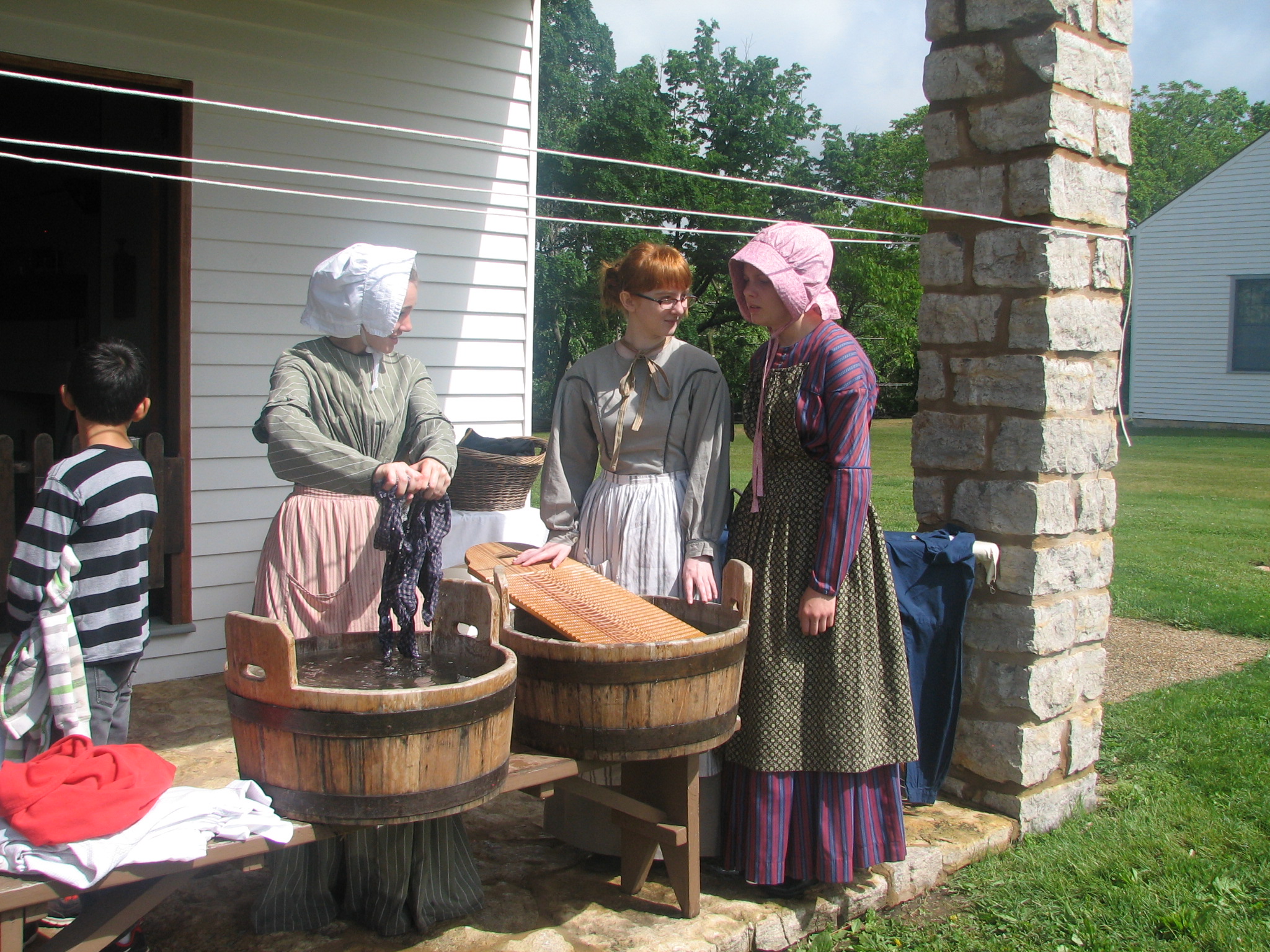 Women dressed as laundresses with laundry buckets and scrub boards stand on a stone porch.