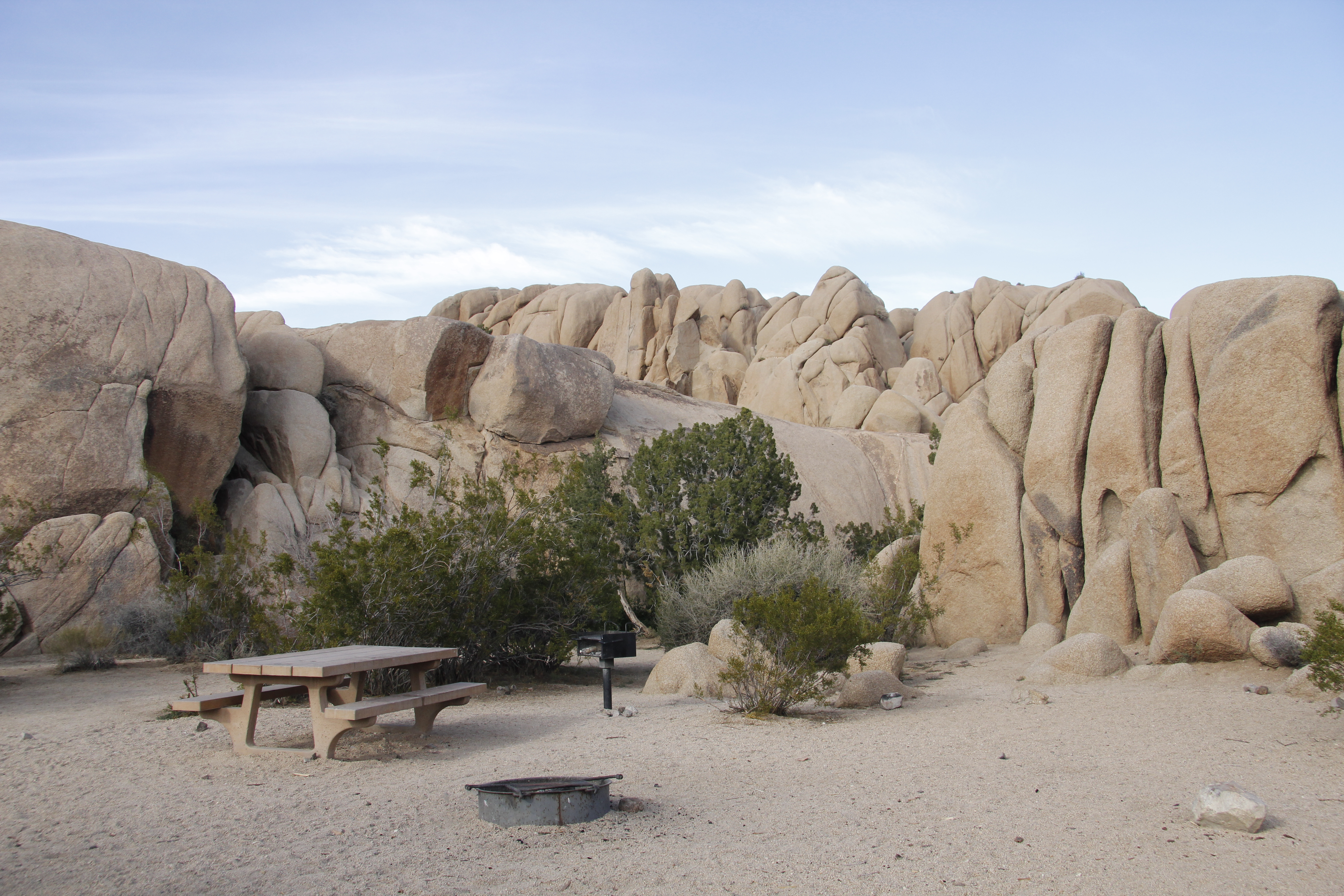 A picnic table and fire pit are in a campsite surrounded by boulders and some vegetation.