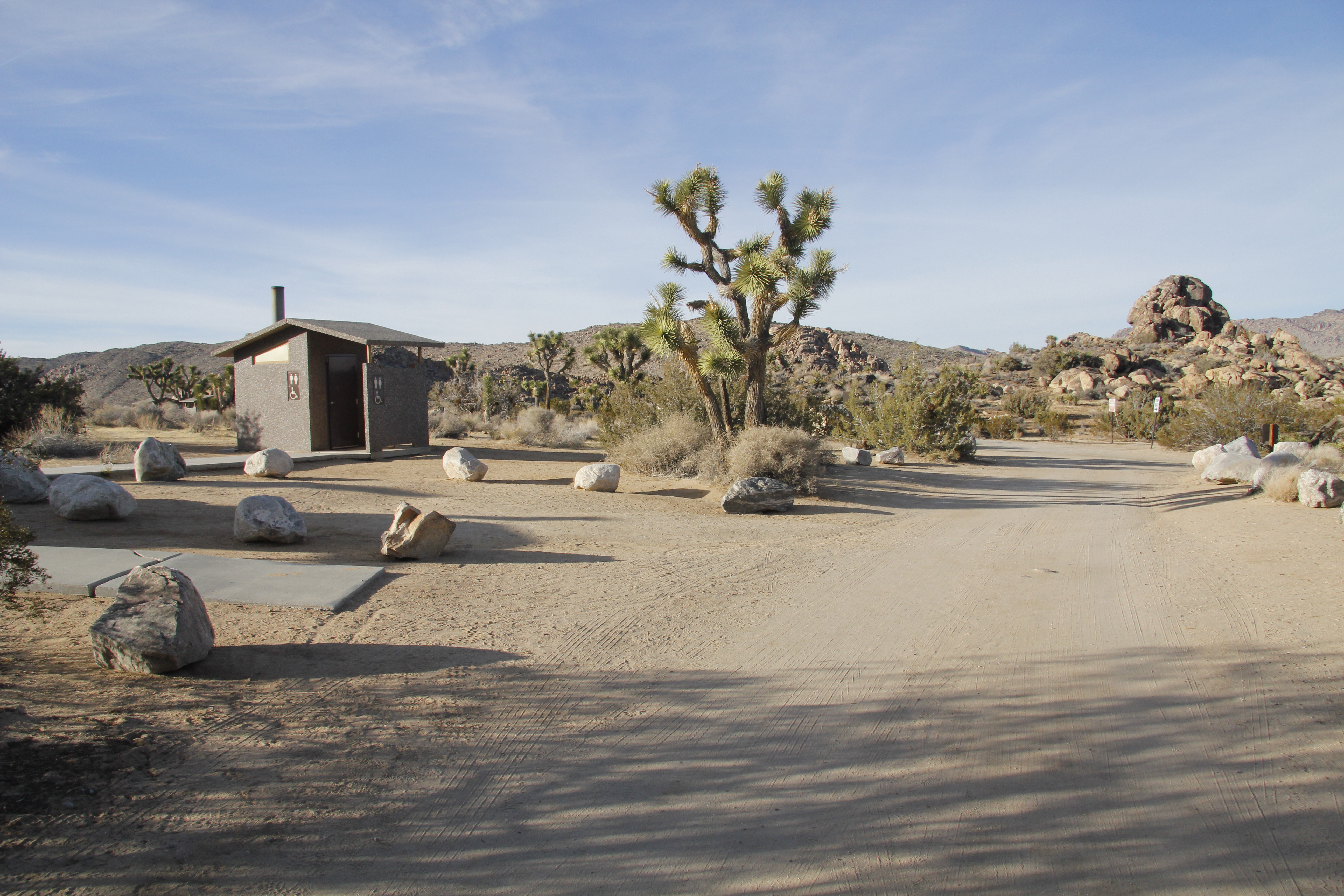 An dirt road and a small paved walking path lead to a vault toilet.