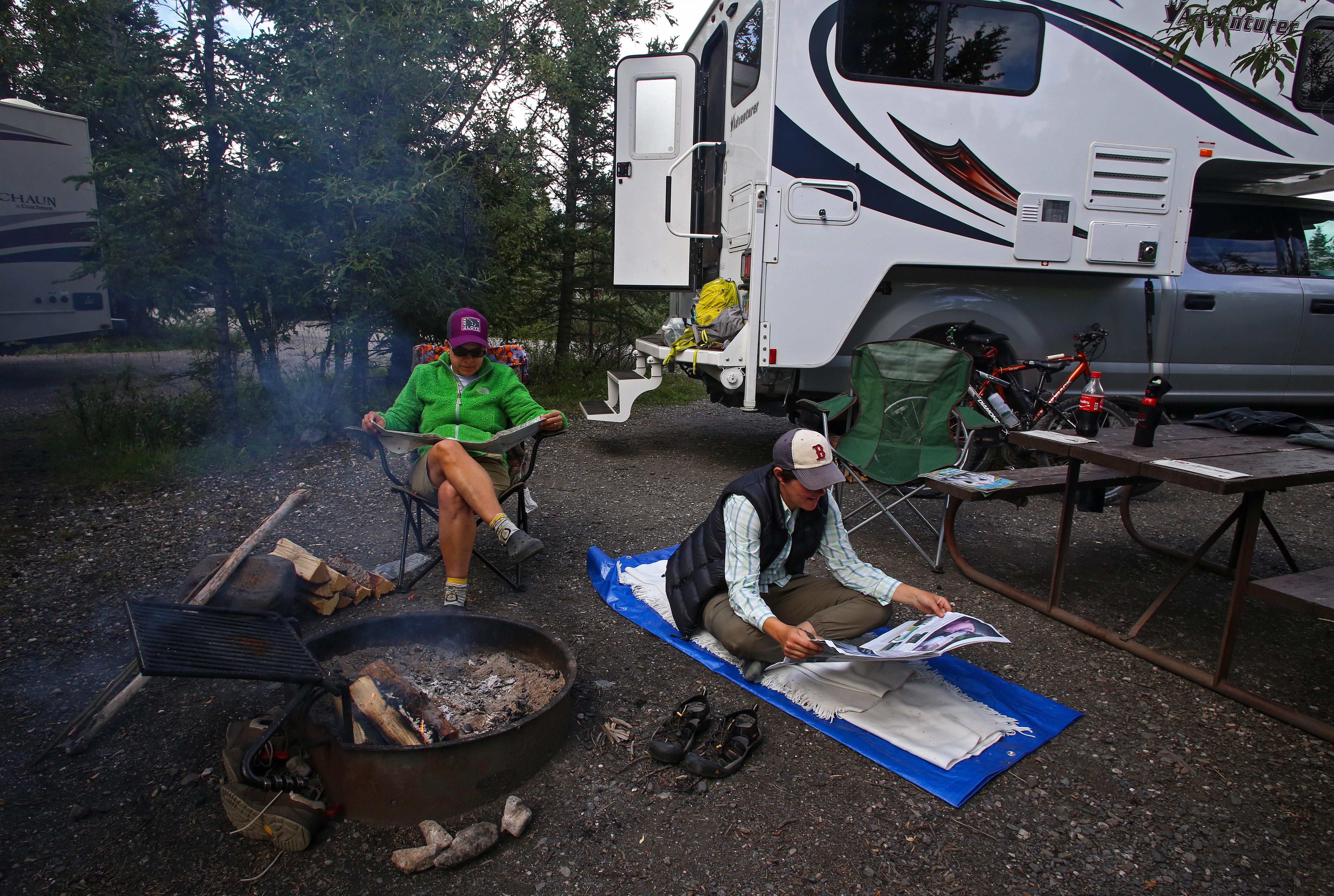 two women sitting near a campfire with a truck and cab-over-camper behind them