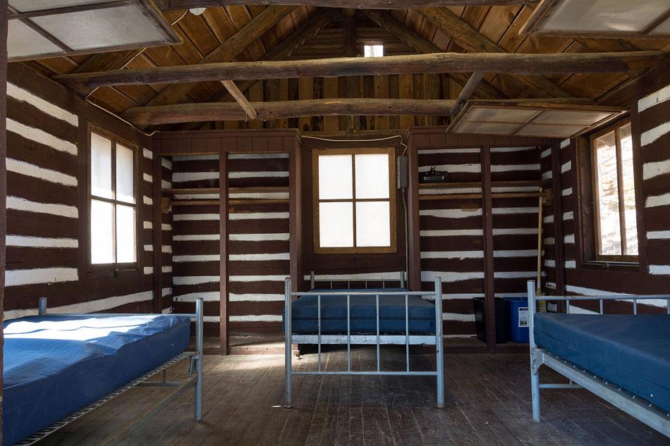 Inside of a log cabin with 3 twin sized bed frames with blue plastic covers.