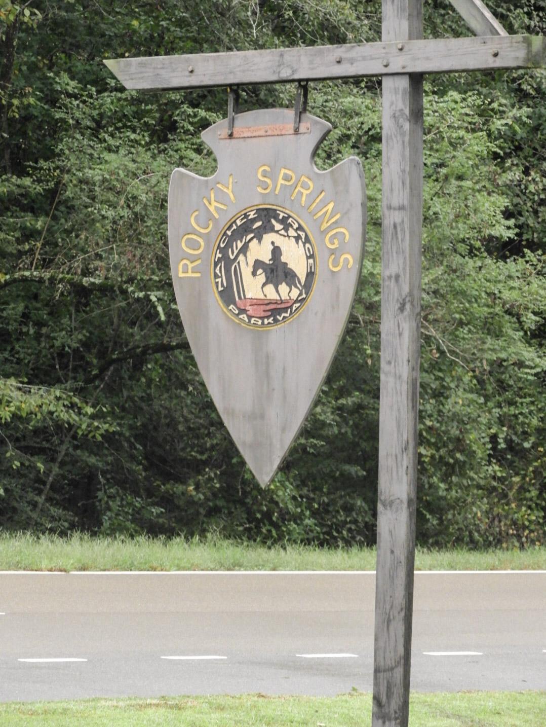 A hanging wooden arrowhead sign with the post rider logo and the words Rocky Springs.