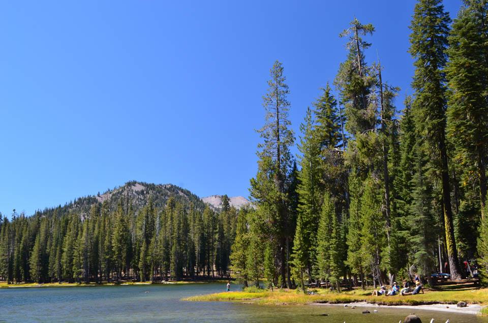 People sit and stand on a grassy shore of a mountain lake.