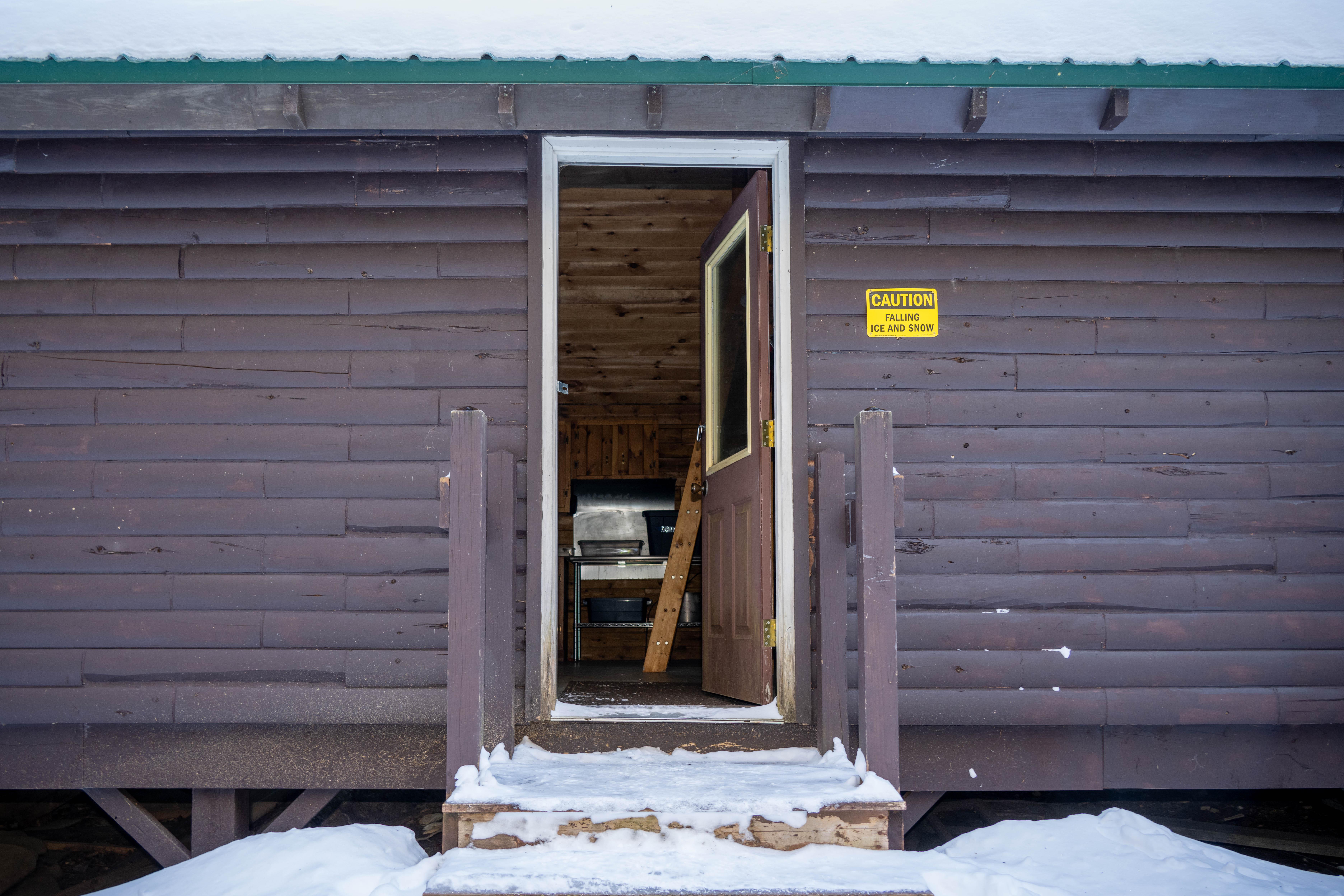 A cropped close up image of an opened door to a brown log cabin. Steps are layered with snow.