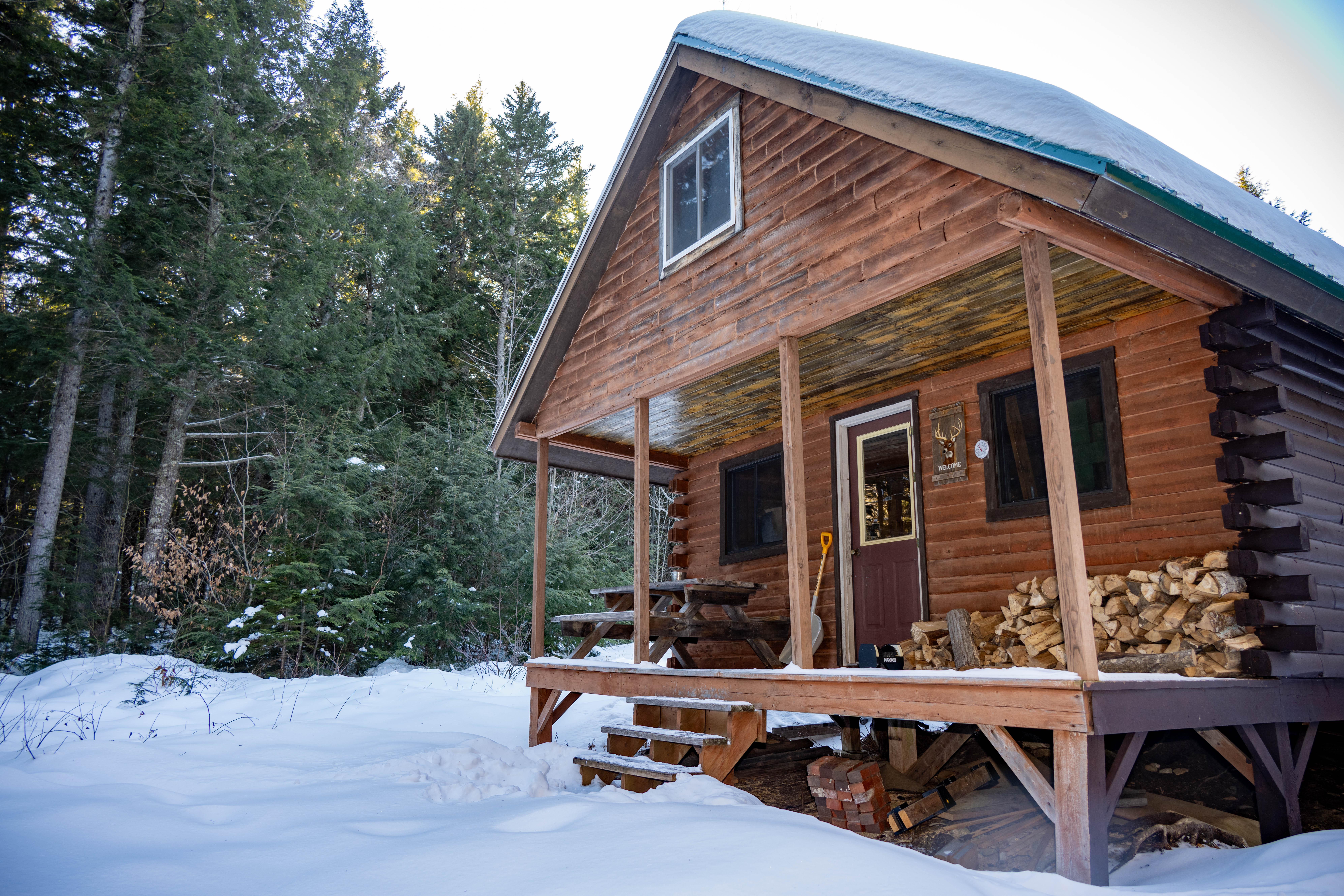 A small wooden cabin with steps and windows is surrounded with thick snow in the woods.