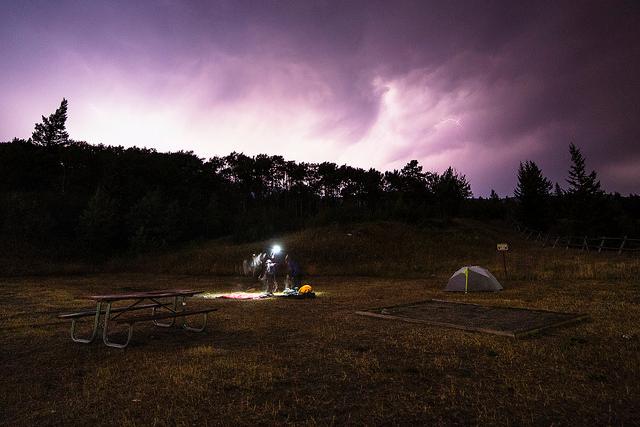 dark cloudy sky over campsite in field with tent, picnic tables, and campers wearing headlamps