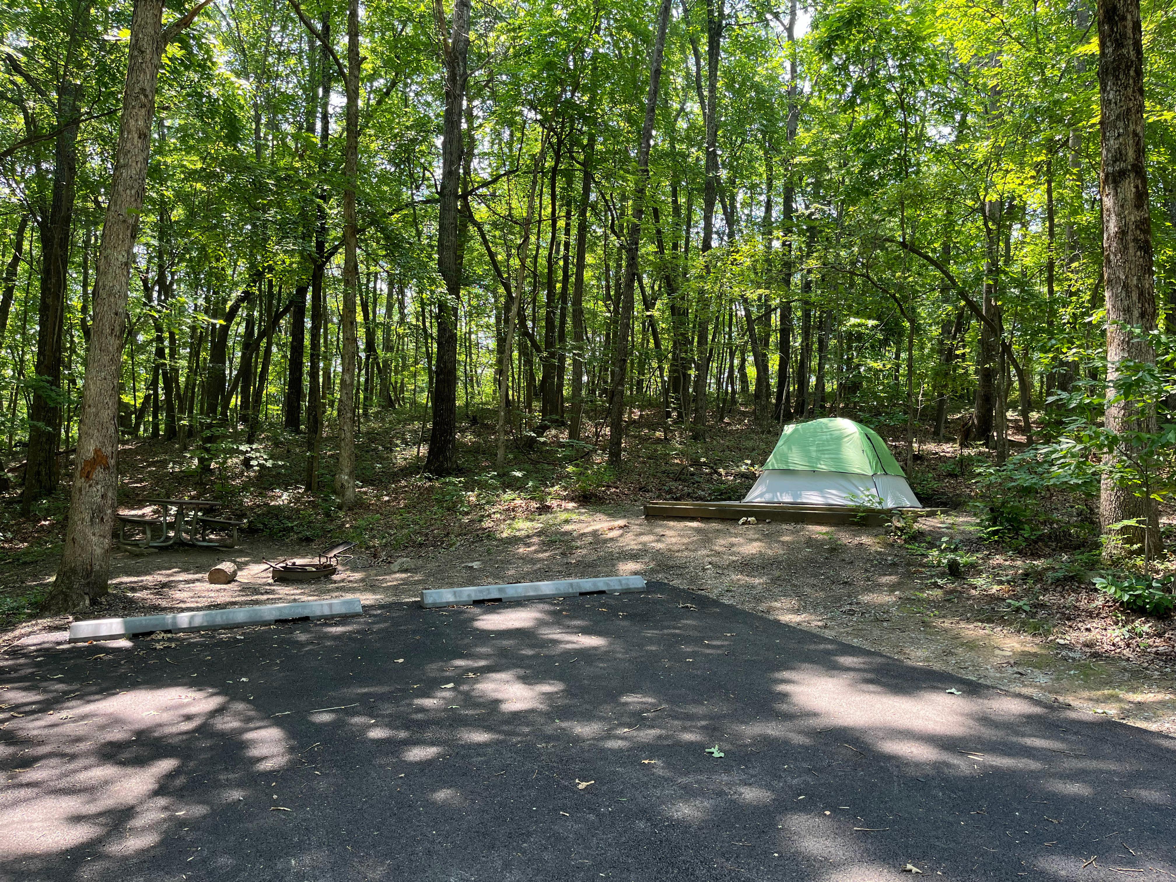 A white tent with a light green rainfly near a picnic table, fire ring, and two paved parking spots.