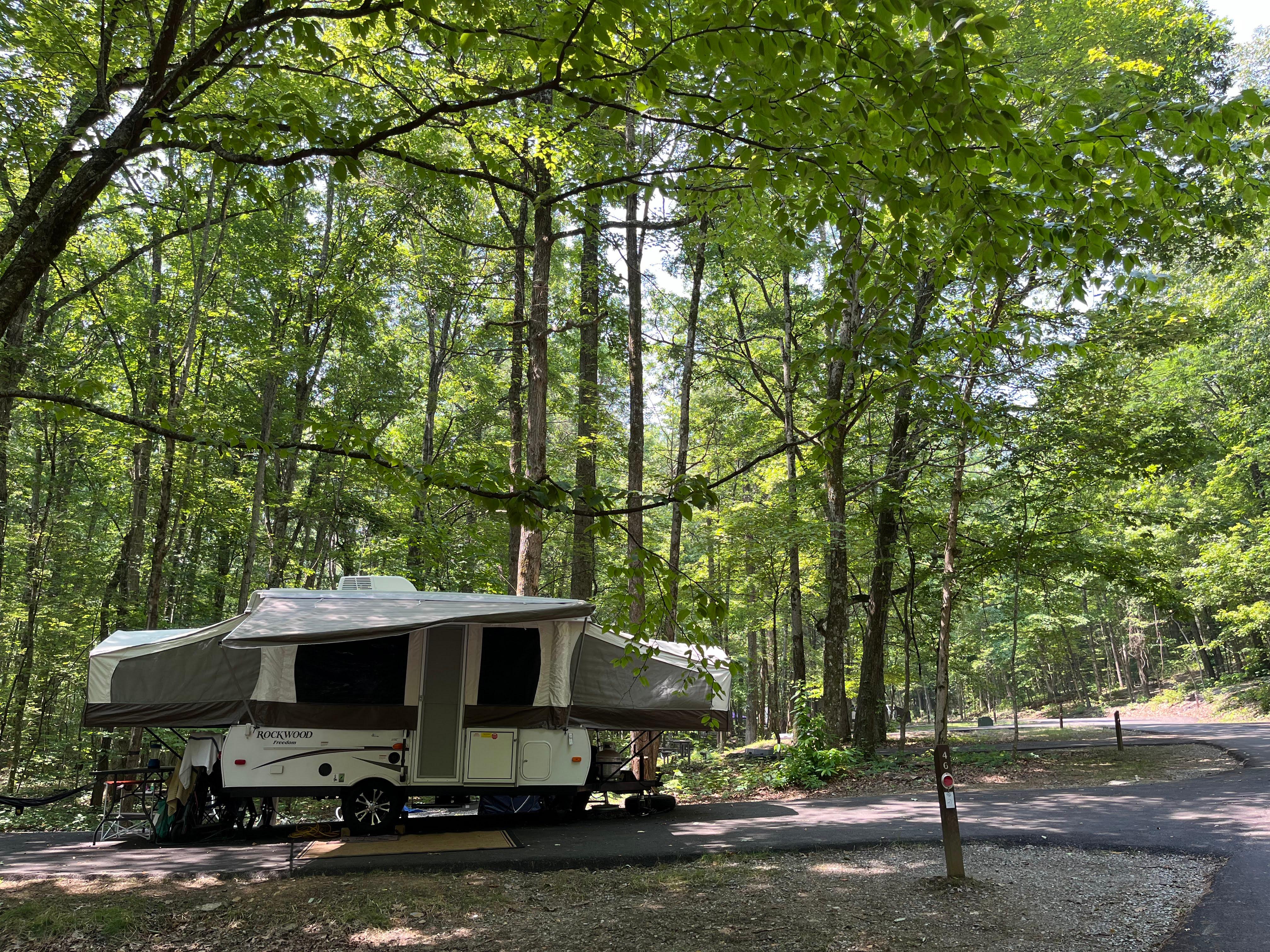 A white pop-up camper shaded by trees, parked in a paved spot beside a wooden post.