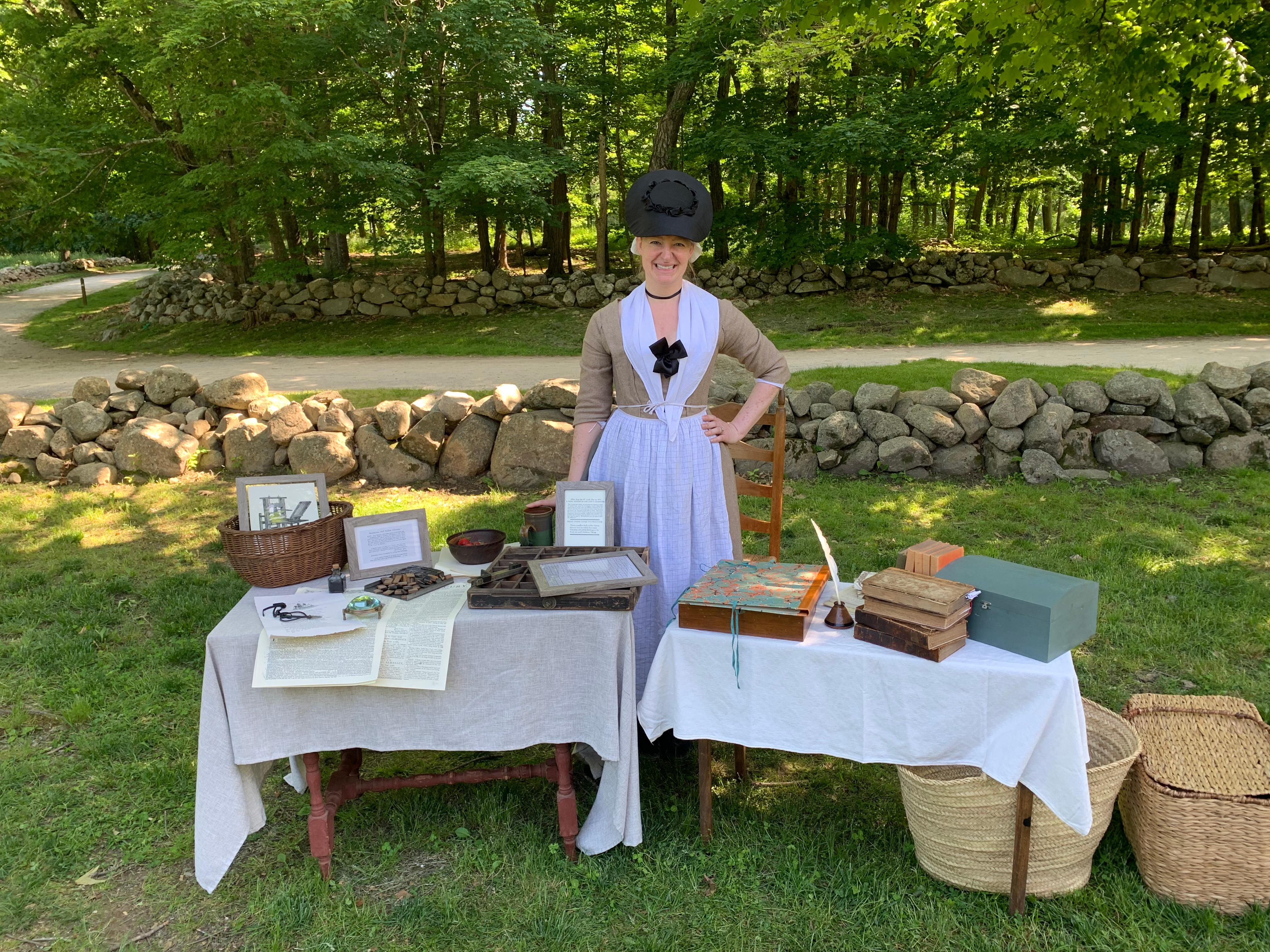 A woman in 1770's clothing stands behind a table filled with materials for printing newspapers.
