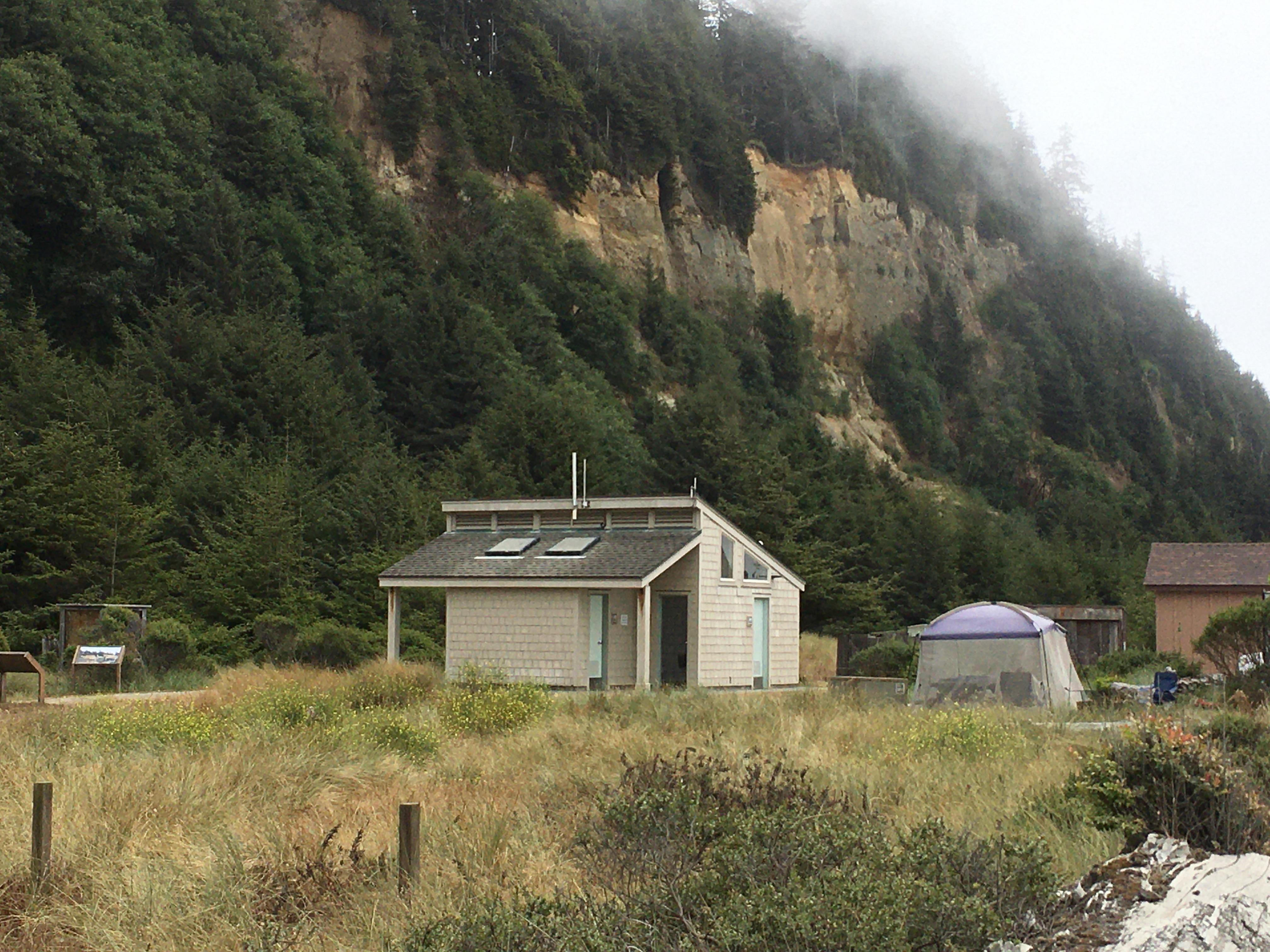 A small building and a tent are seen in front of a steep bluff.