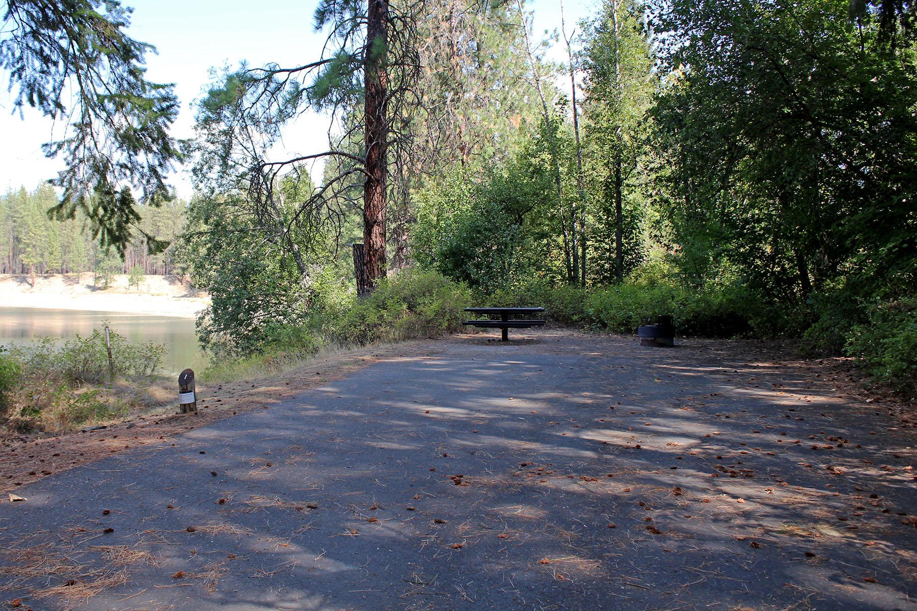 a campsite with a large paved parking area, accessible picnic table, and fire pit, with view of lake