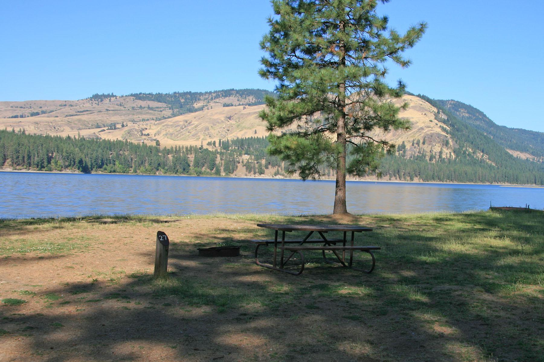 picnic table and fire pit next to campsite number post, with lake in background