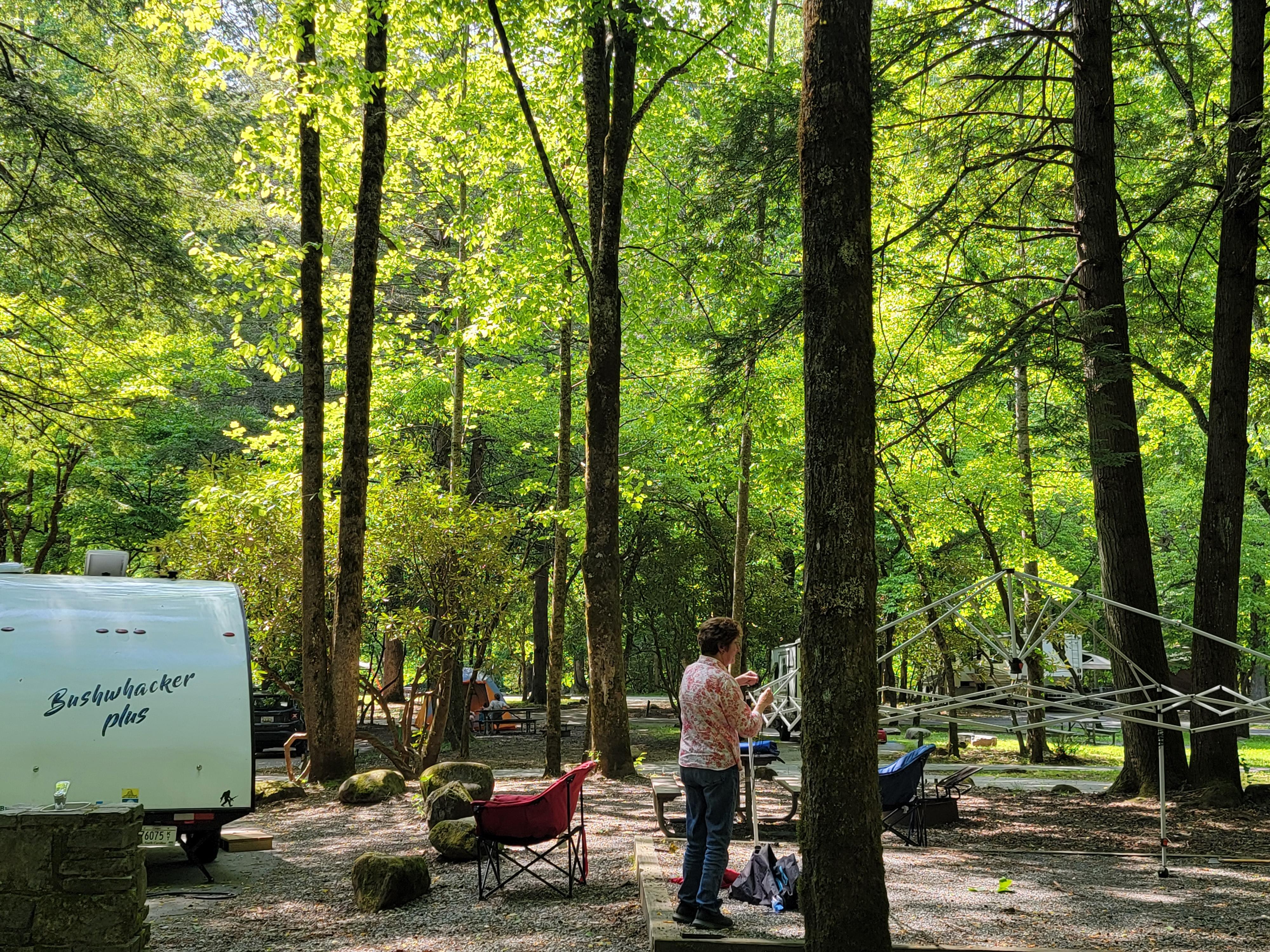 A person standing on a level gravel area setting up a canopy tent, all next to a white trailer.