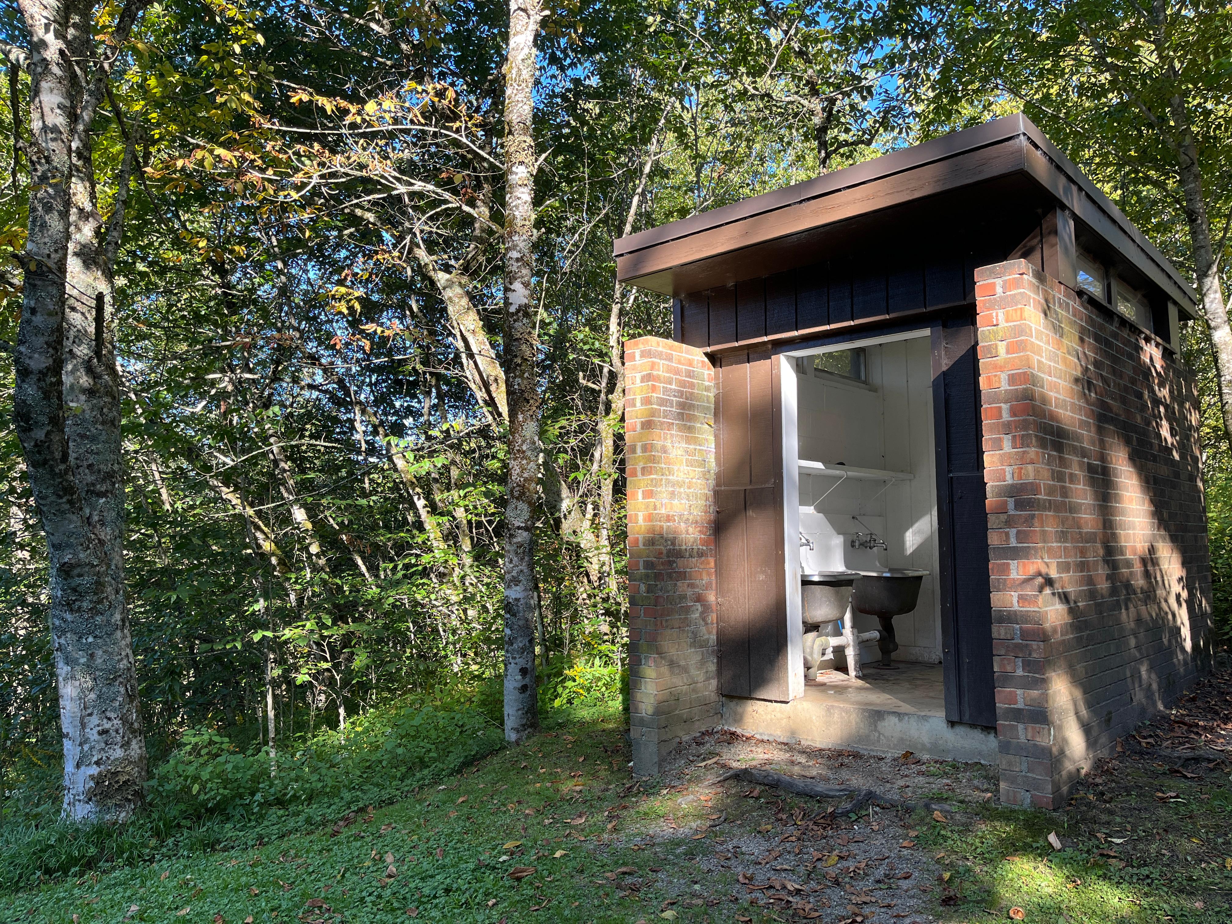 A brick structure with a brown roof and small windows surrounded by trees. Two sinks visible inside.