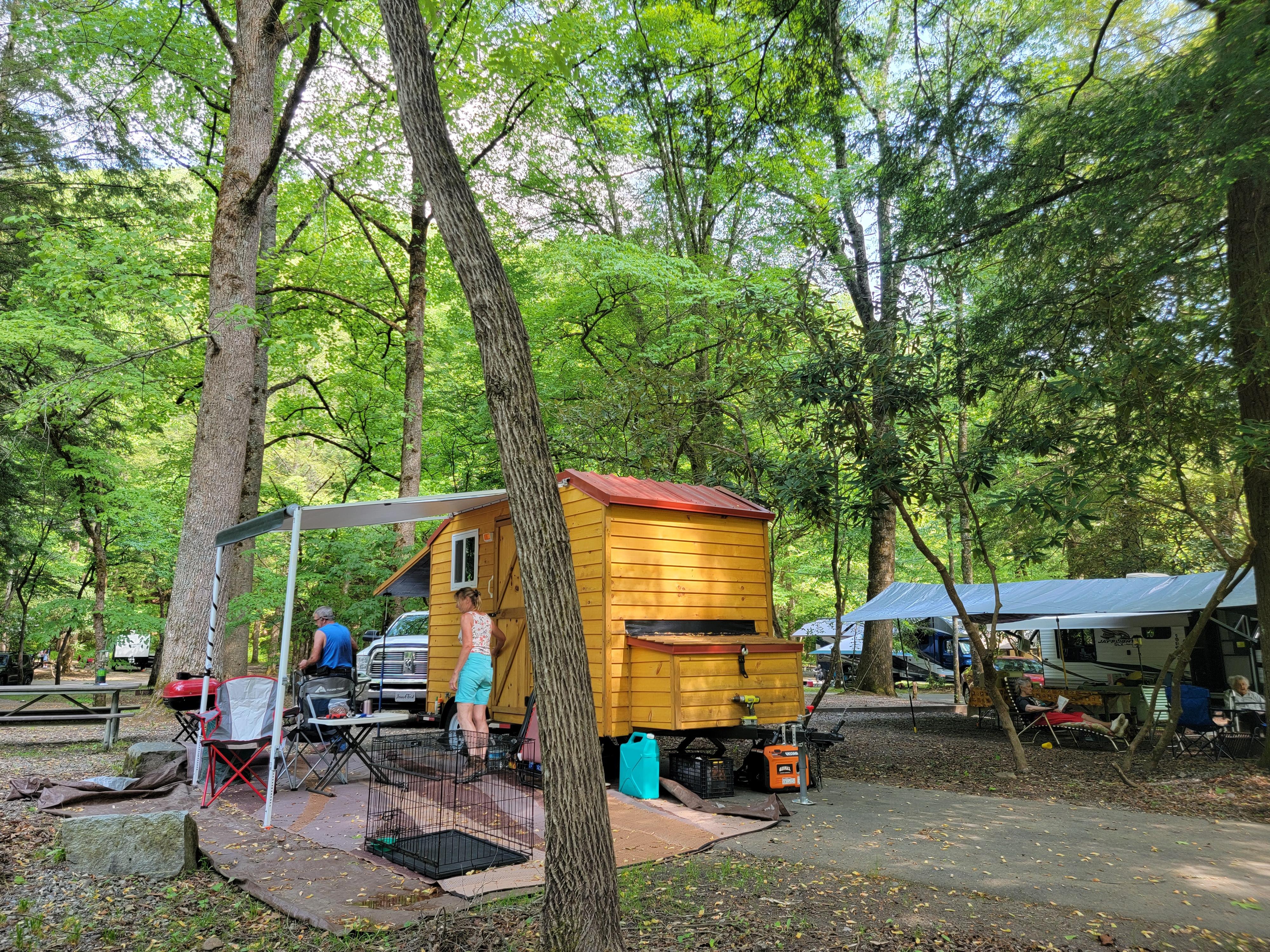 A trailer that resembles a tiny home with a red roof and porch cover parked in a campsite.