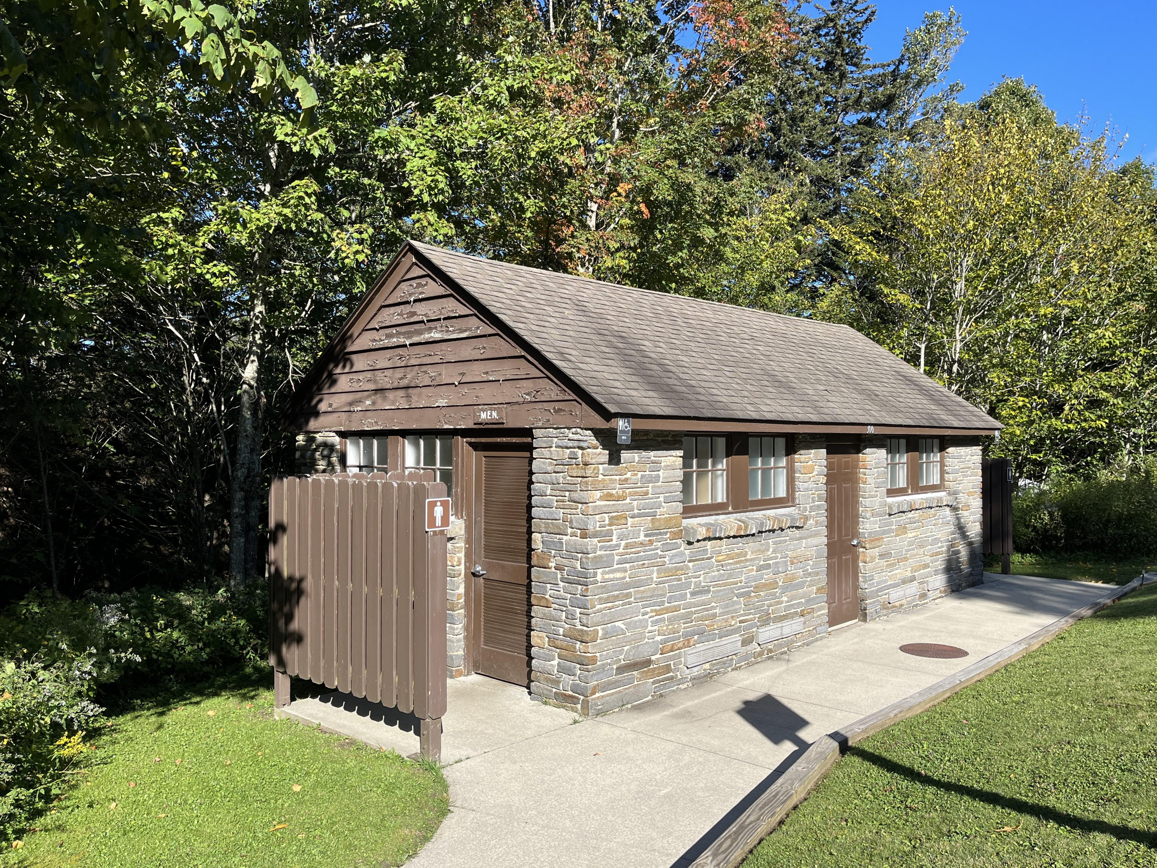 A paved sidewalk leads to a brick restroom building with a brown roof and trees in background.