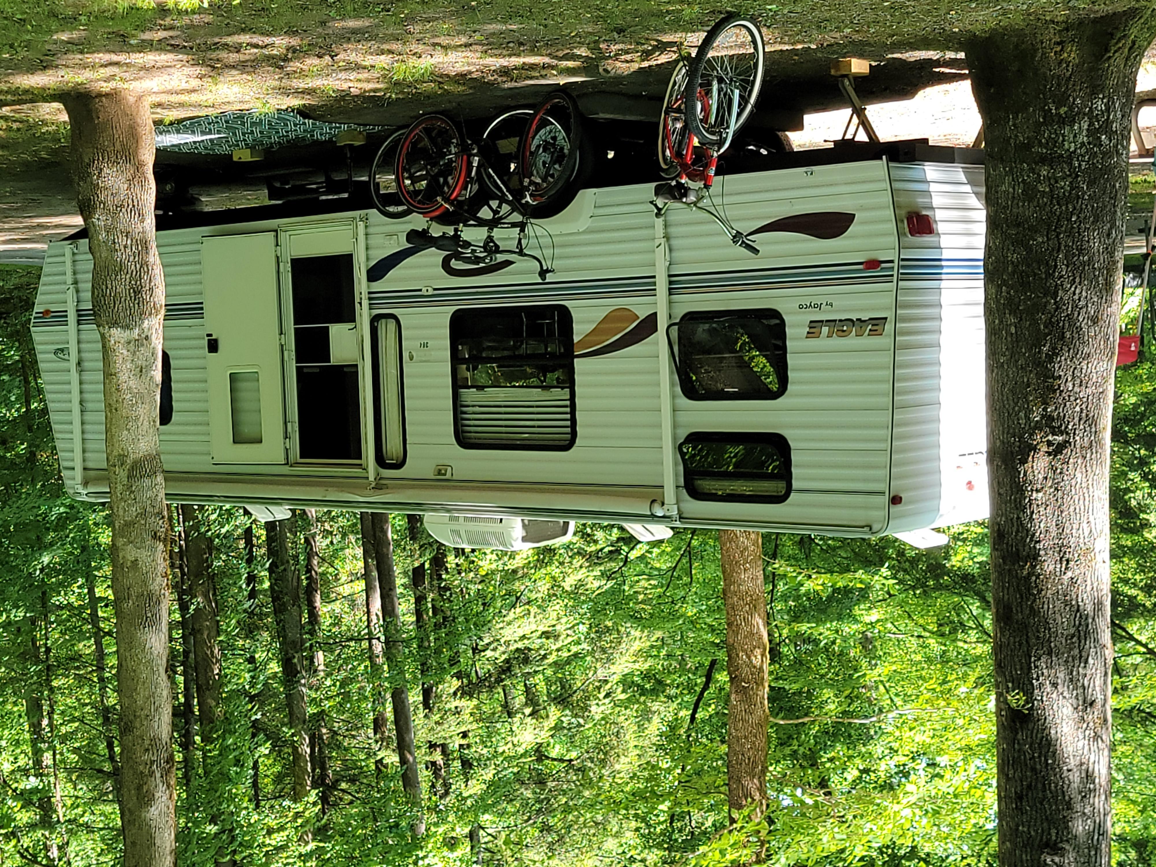A white camper trailer with multiple bikes parked on the side, all under the shade of trees.