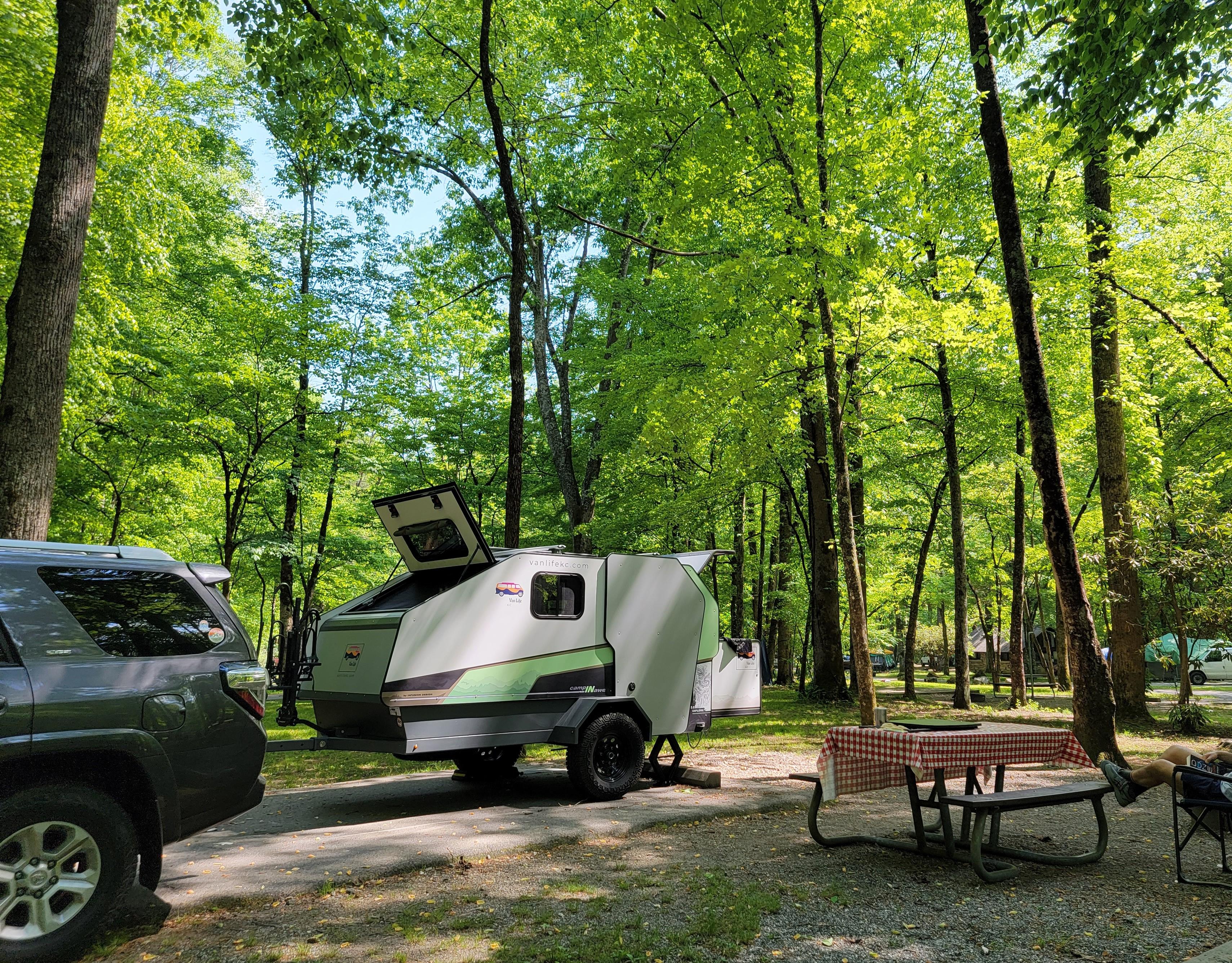 A white trailer and gray SUV parked at a campsite with a red checkered cloth on its picnic table.