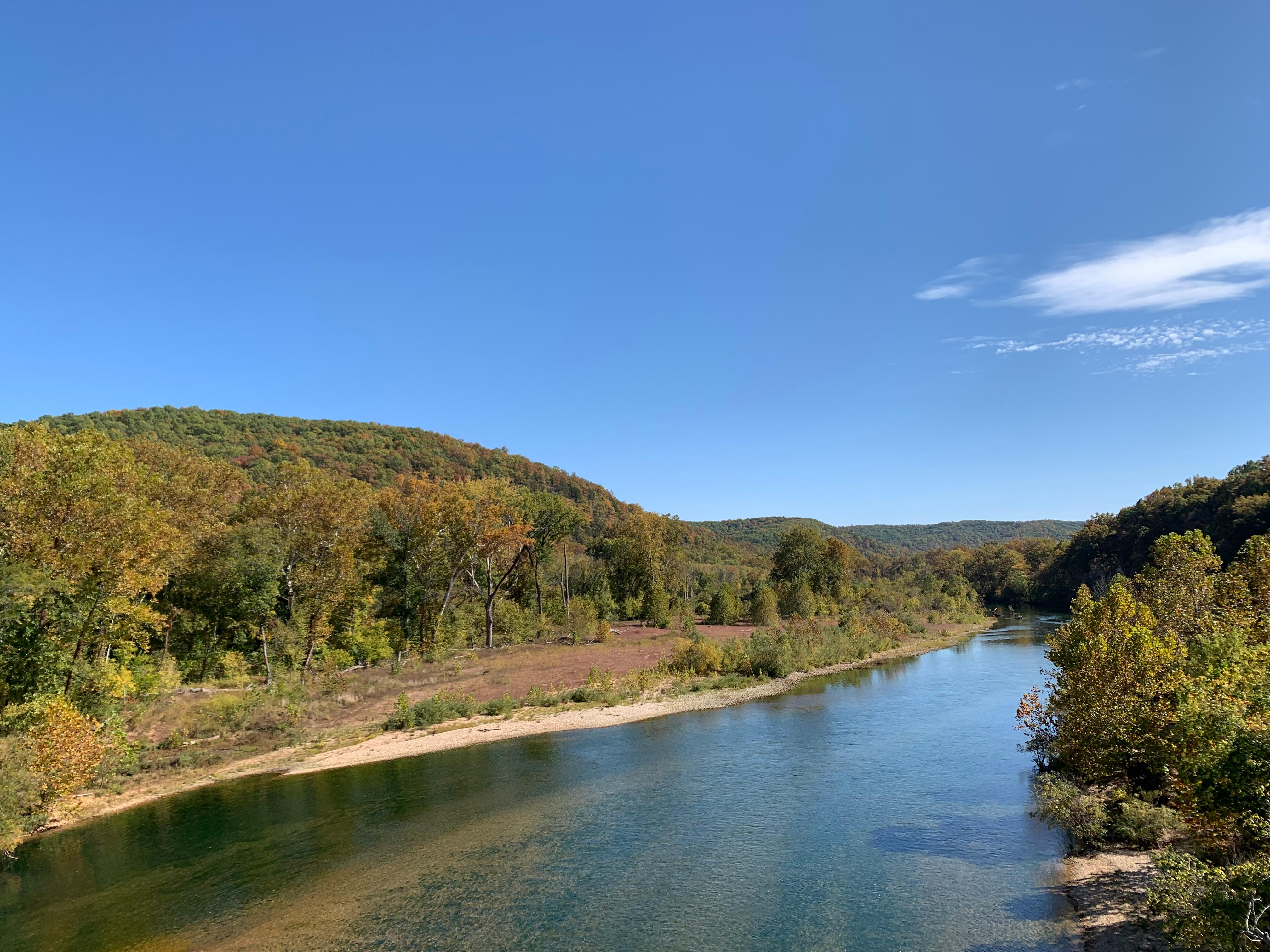 A blue river flows along a large gravel bar, with various plants and trees.