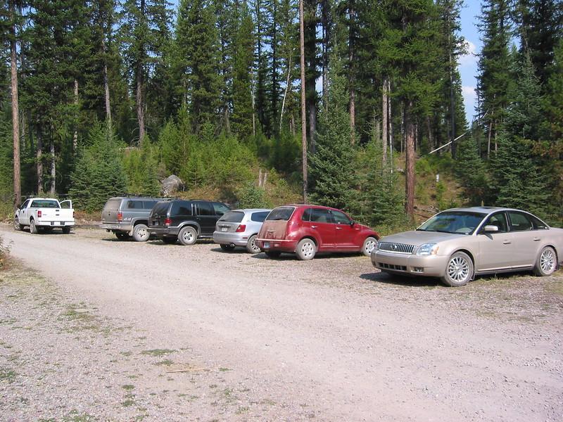 Six dusty cars are parked along side a dirt road with a forest in the background on a sunny day.