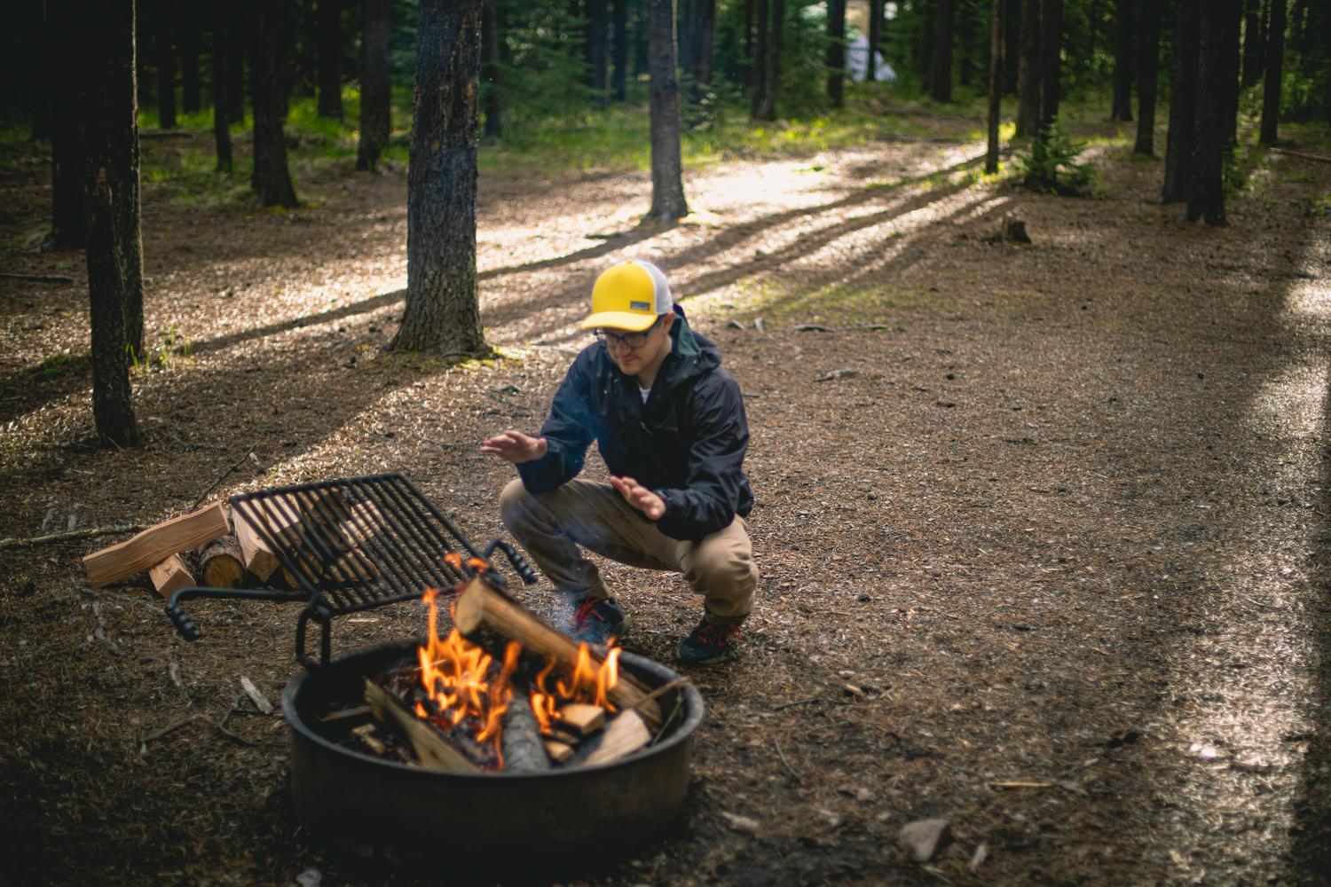 A camper wearing a bright hat warms their hands near a campfire in a forest clearing.