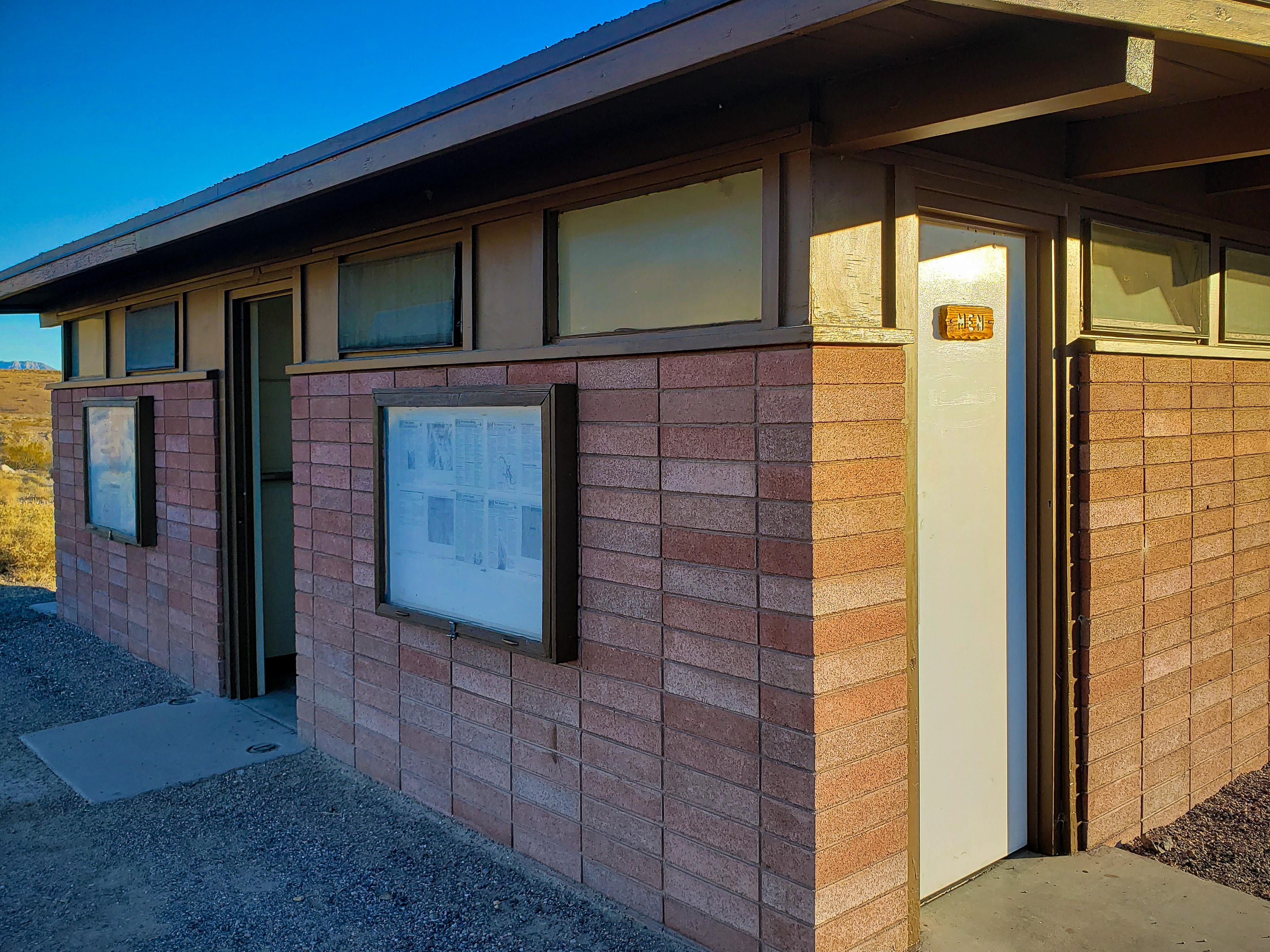 2 doors with square concrete ground pads on red brick building. 1 reads Men, other is open.