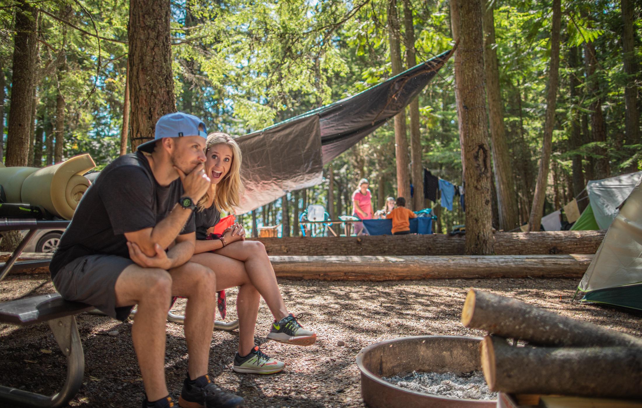 Two people sit by a campfire ring in a forested campground with tents and other people in the backgr