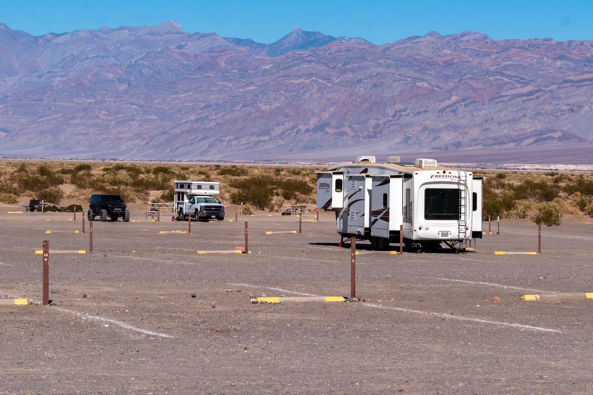Large mountains in the distance fill the background of an open, gravel lot with 2 RVs.