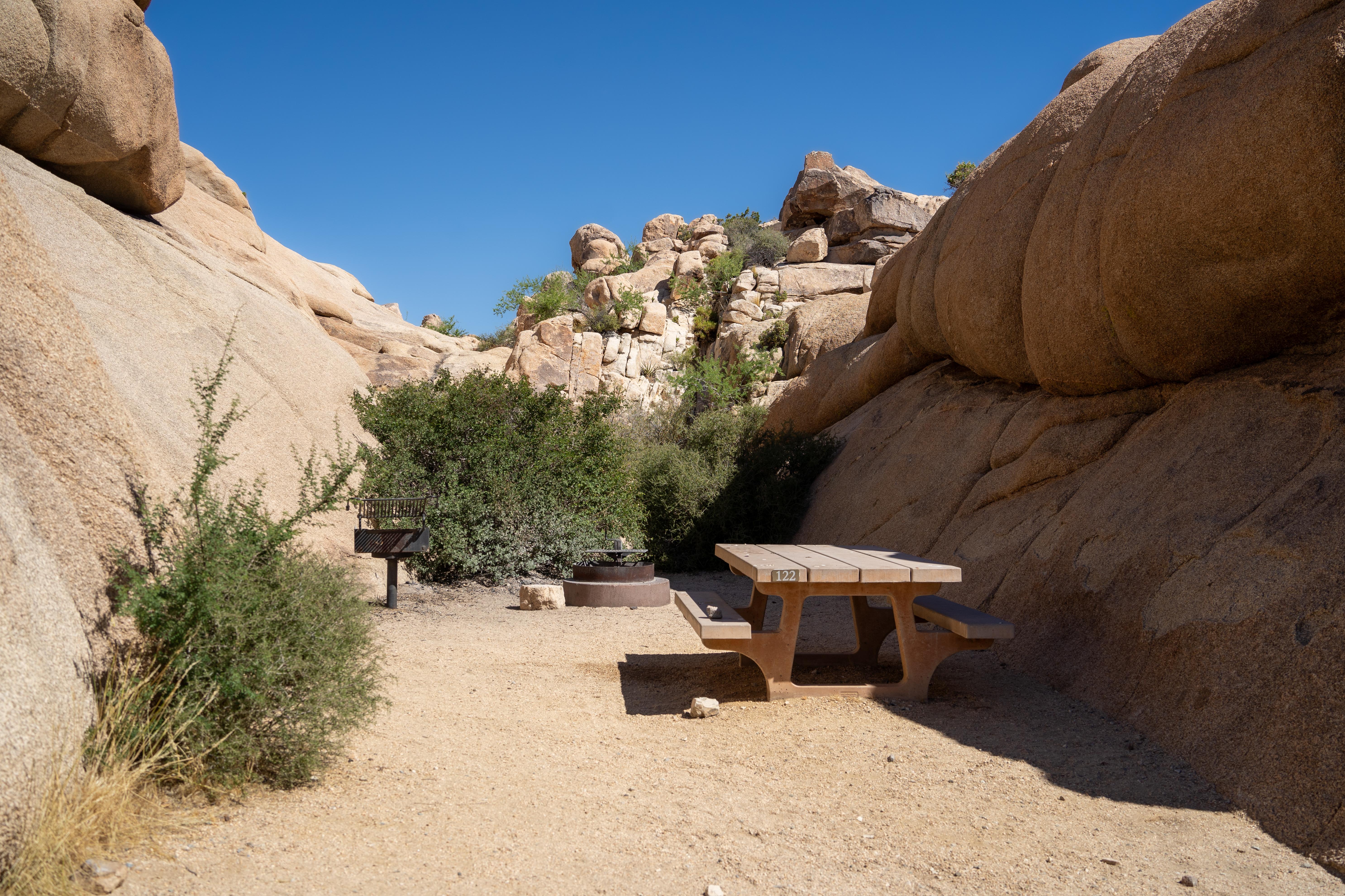 A picnic table, grill, and fire pit sit in a sandy area between large rock formations.