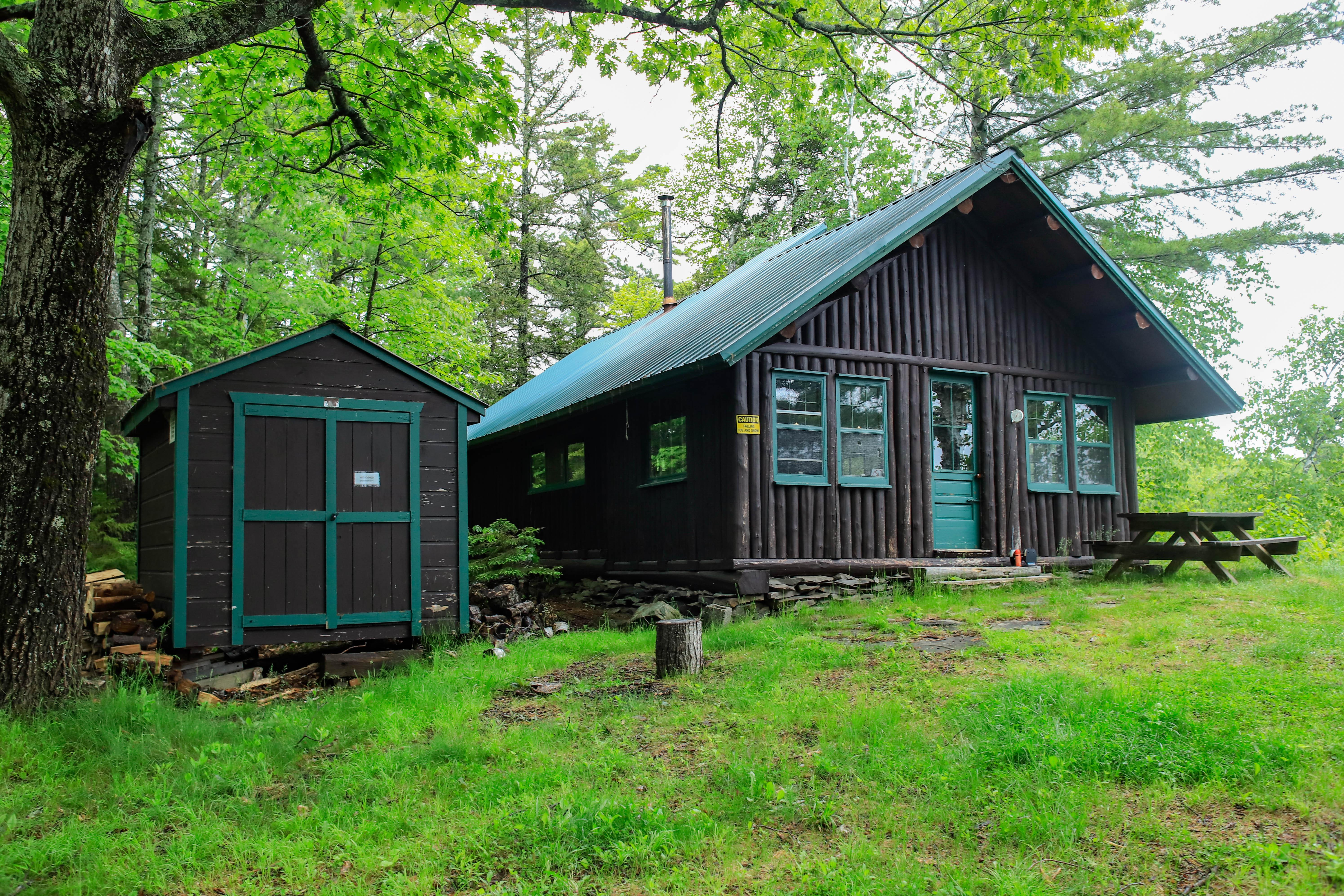 A dark brown log cabin next to a woodshed in the woods