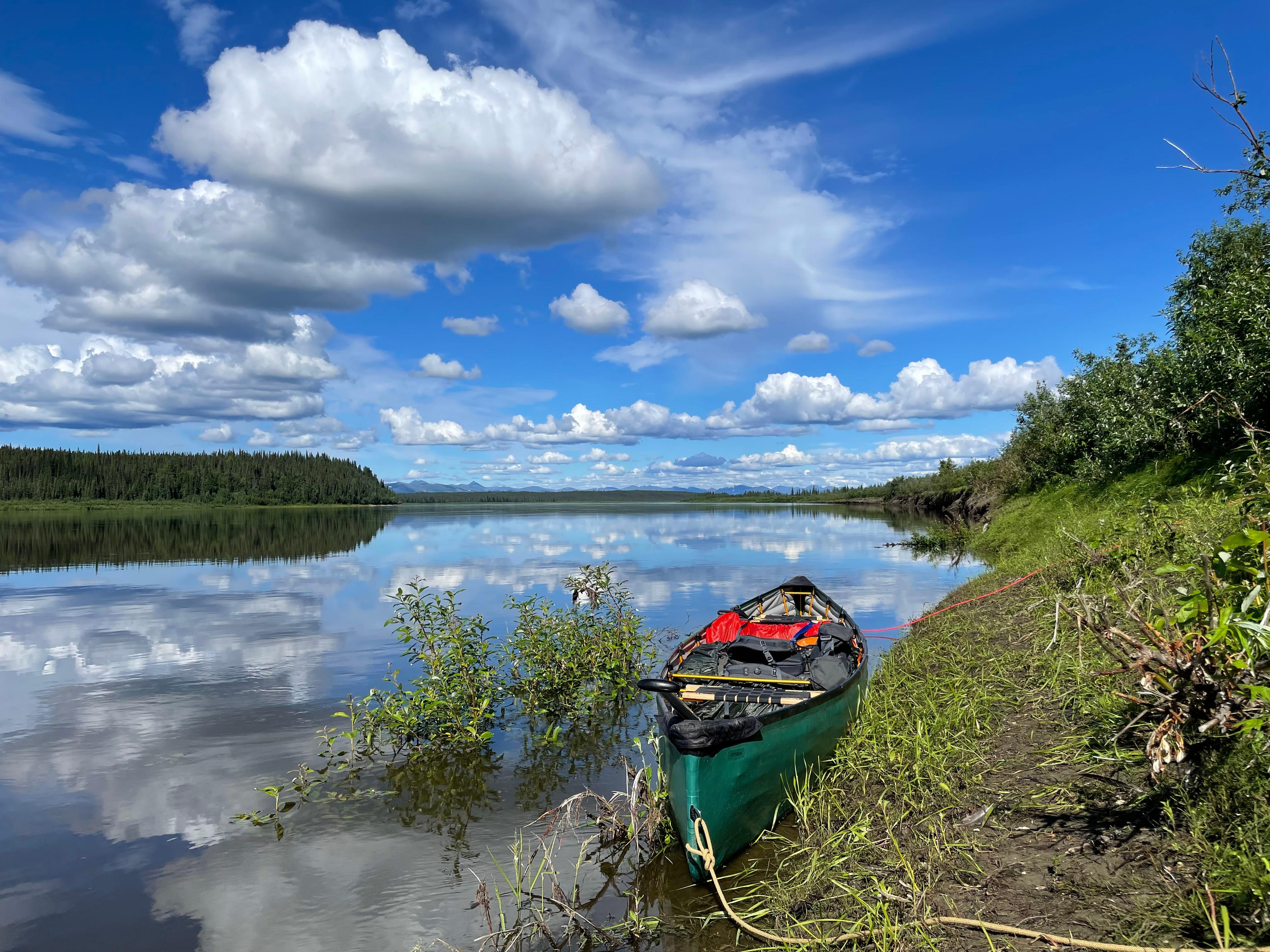 A green pack canoe sits on the bank of the Kobuk River. Clouds are mirrored on the water's surface.