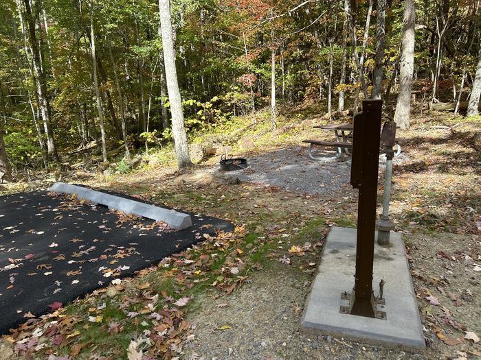 A campsite in the forest with a gravel pad, fire ring, and hookups in view beside the parking spot.