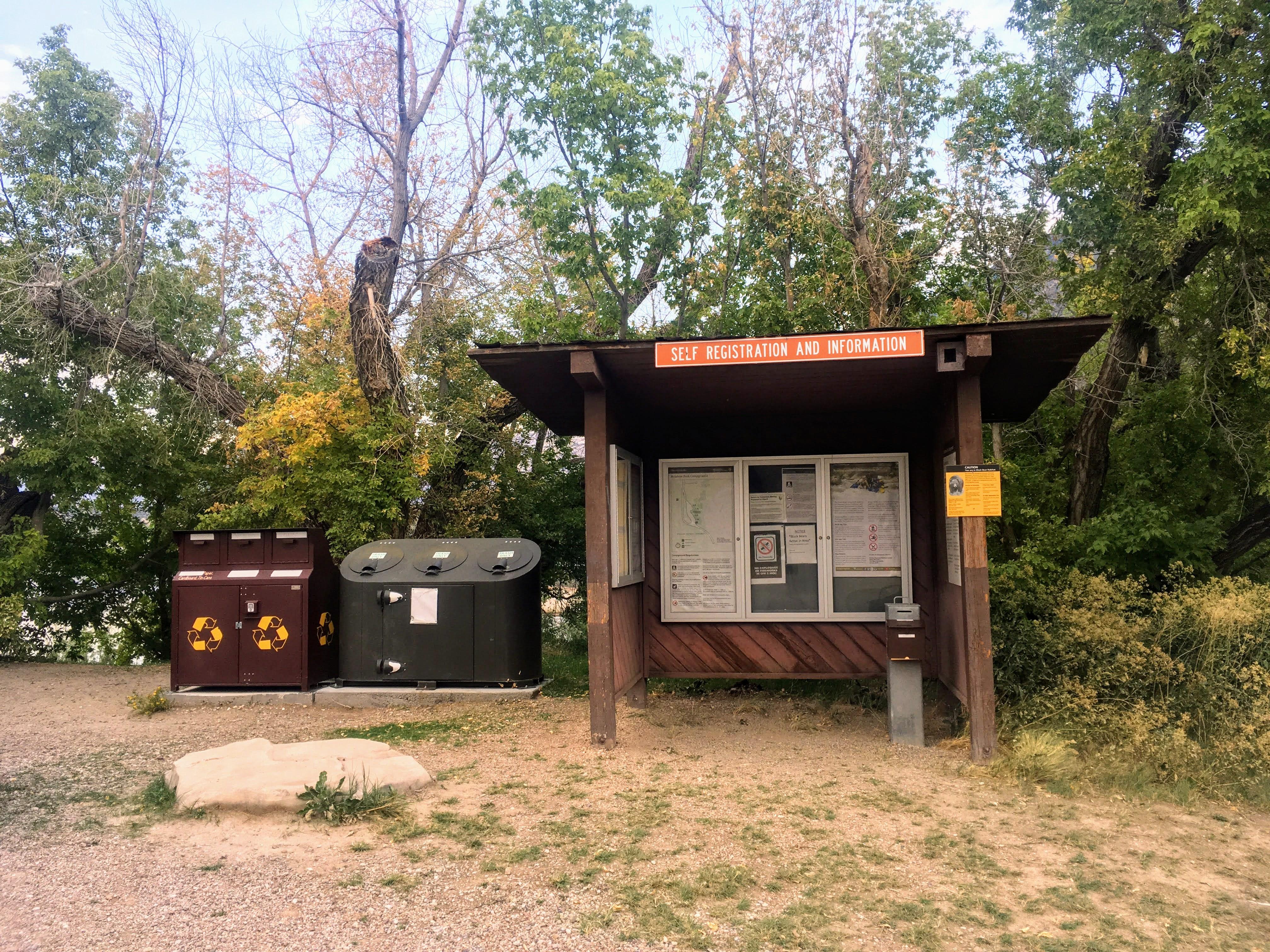 A shaded wooden kiosk with park information beside some trash receptacles.