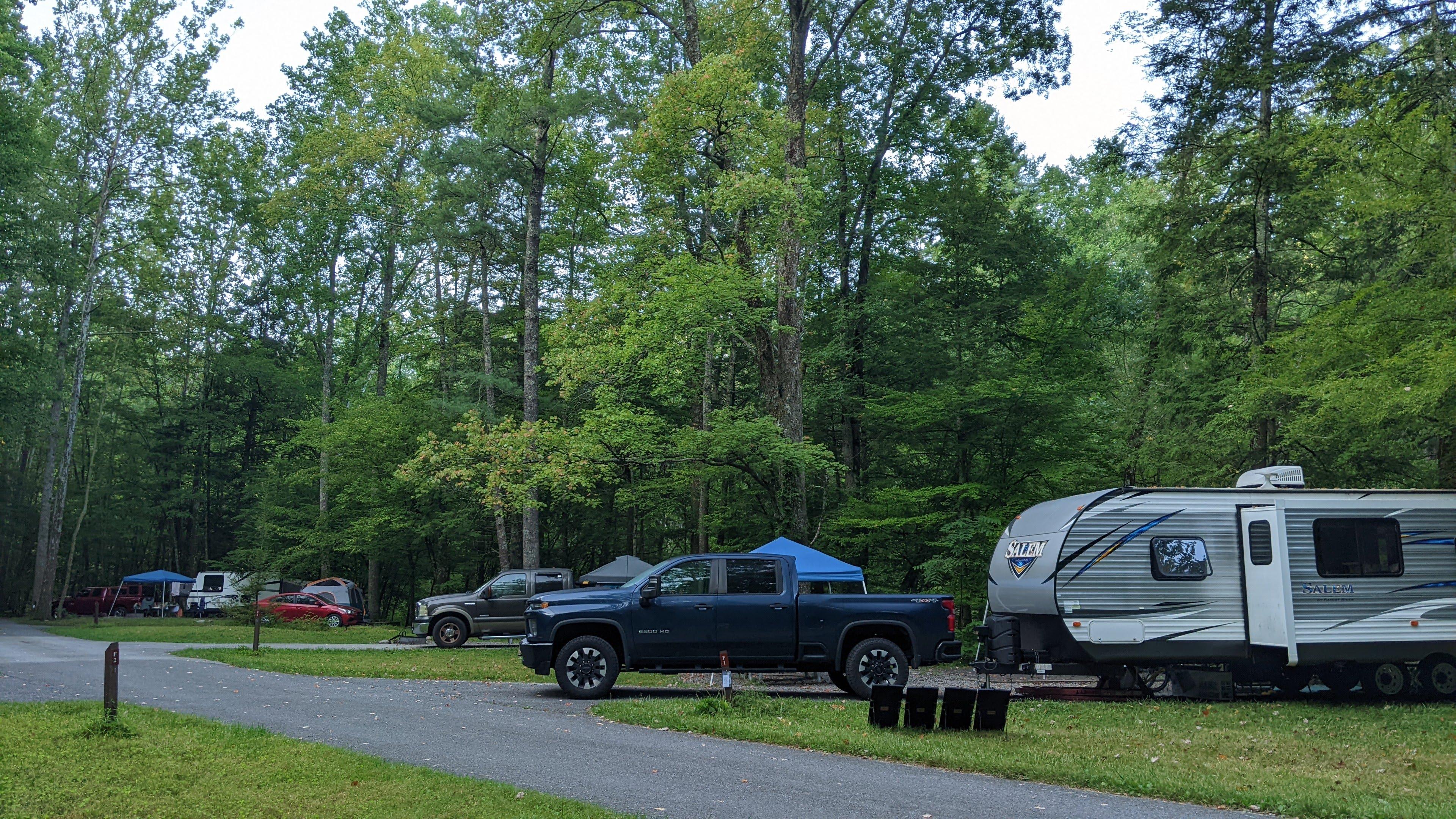 A gray and white RV attached to a blue truck. Other sites and trees are in the background.