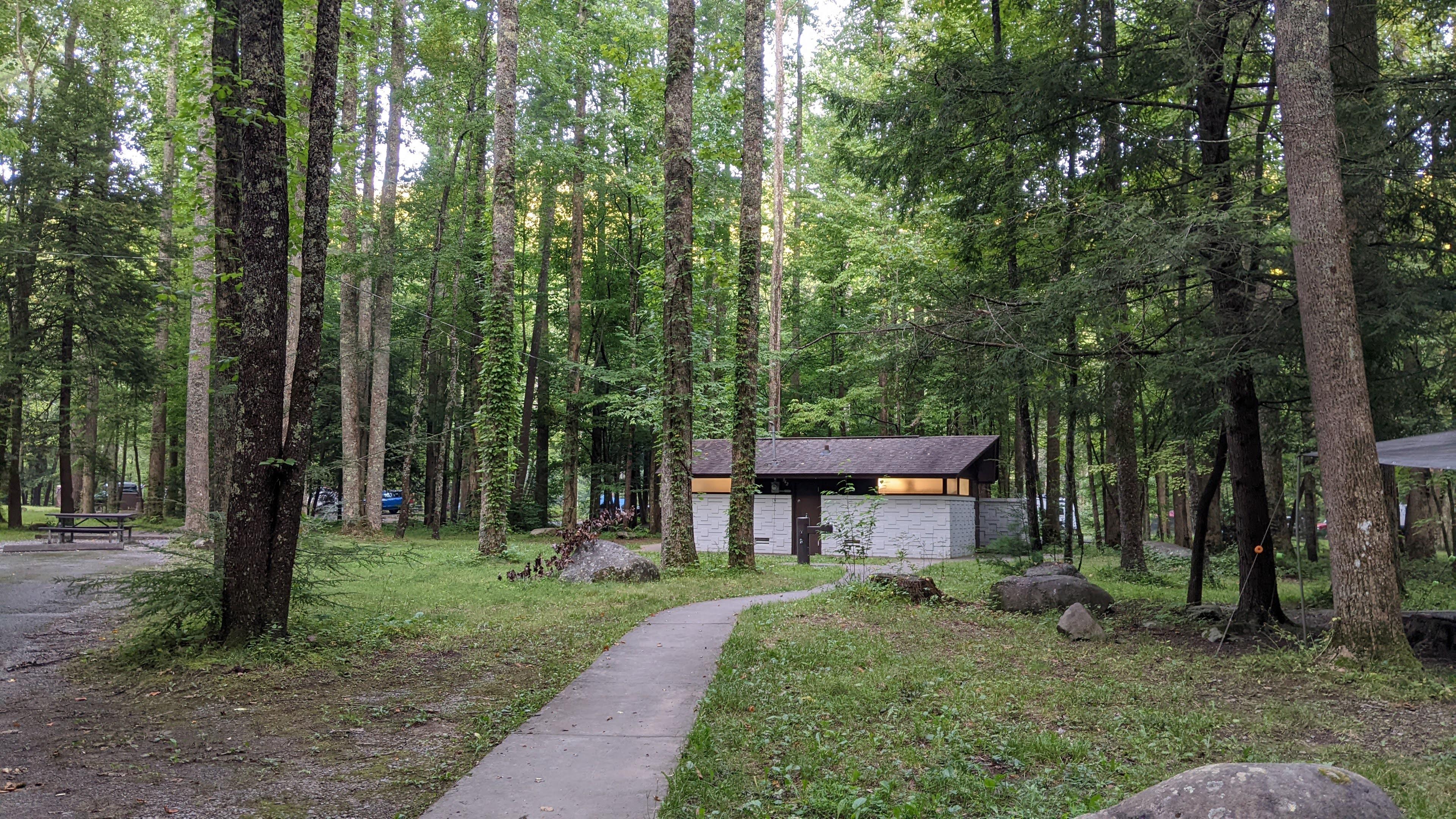 A paved sidewalk leading to a restroom building with a brown roof. Trees and grass surround it.