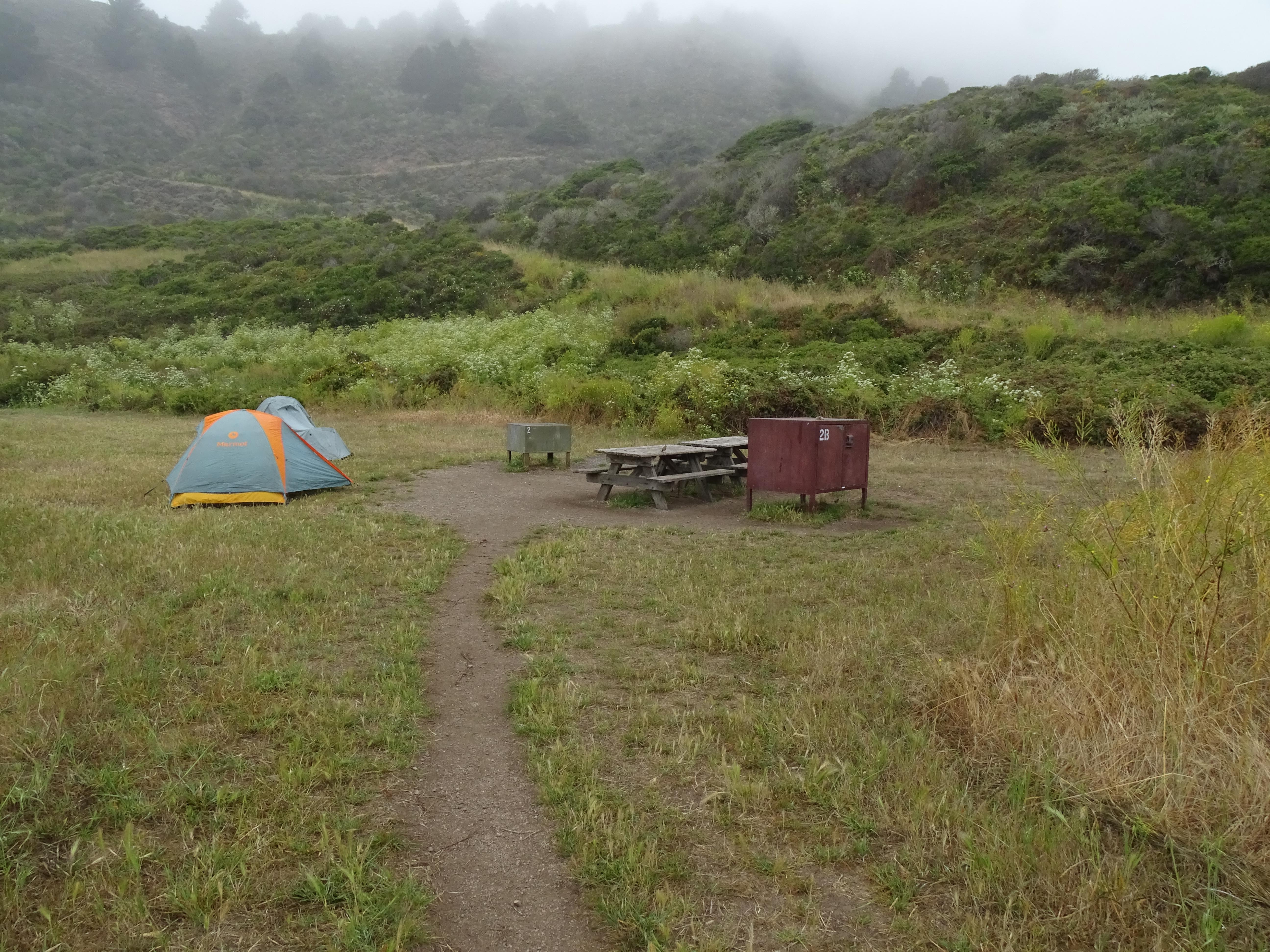 A campsite in a meadow at the base of a hill containing two picnic tables, two food storage lockers,